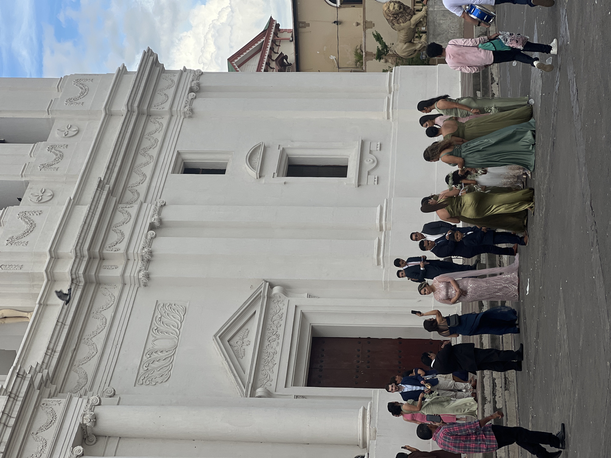 This image depicts an outdoor scene in front of a large, white, ornate building, likely a cathedral or historic church, located in León, Nicaragua. The architecture features classical columns, intricate carvings, and a large wooden double door. A group of people, dressed in formal attire, are gathered on the steps and the paved area in front of the building. Several individuals are holding smartphones, suggesting they are taking pictures or videos of an event, possibly a wedding or special ceremony.
In the foreground, a man in a plaid shirt walks away from the camera. To his left, a group of women in formal dresses, some in shades of green and pink, are standing together. Some of these women appear to be bridesmaids, and there are also young girls in white dresses, possibly flower girls. To the right of this group, a man in a suit is visible, along with other guests.
In the background, behind the main building, another building with a red and white decorative roofline is visible, along with a statue of a lion. The sky above is partly cloudy. The overall atmosphere suggests a social gathering or event taking place at a prominent landmark.