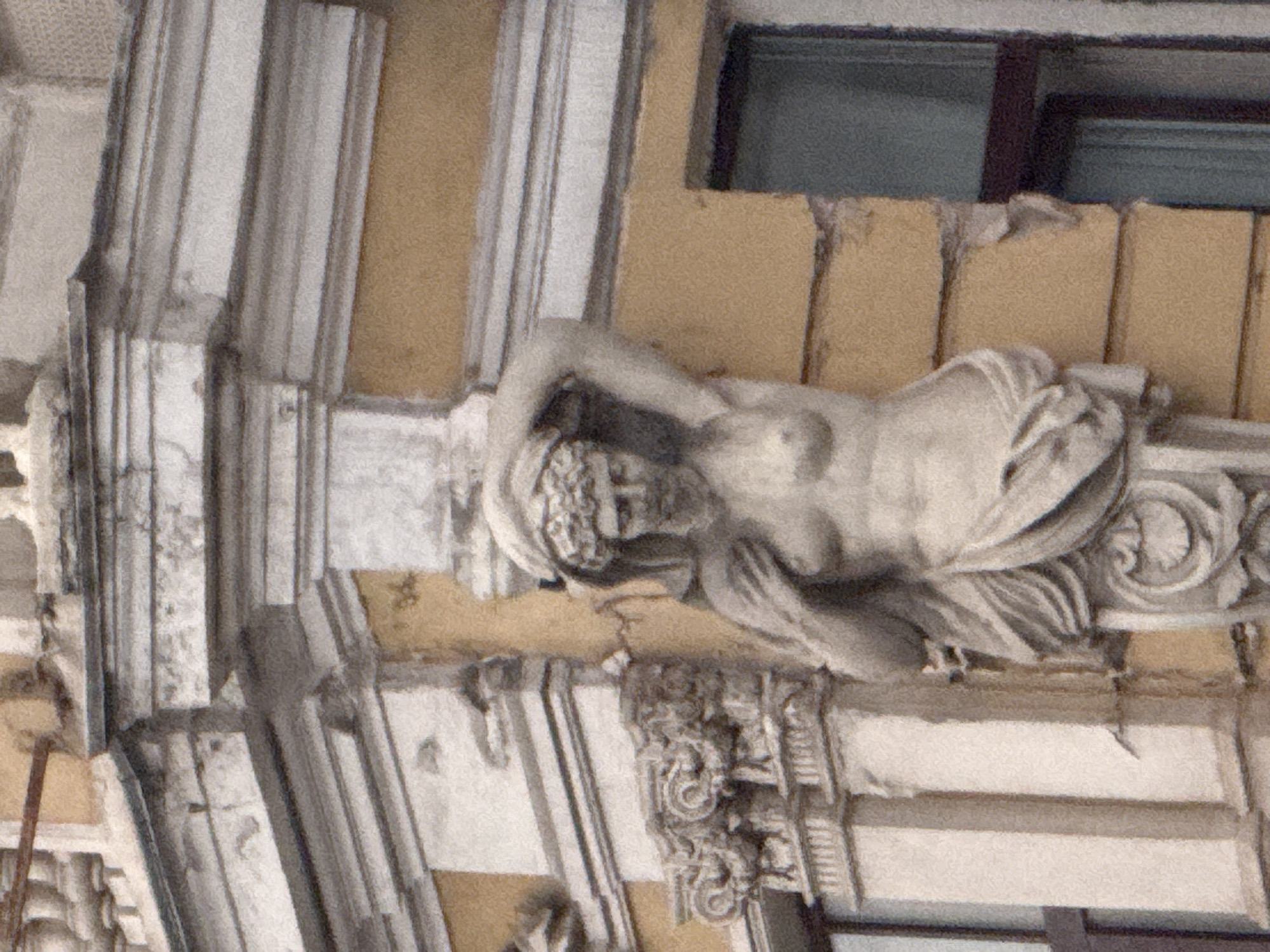 A stone statue of a nude female figure is affixed to the exterior corner of a building. The statue is partially draped with a cloth that covers her lower body and wraps around her waist. Her left arm is raised above her head, and her right arm rests at her side. The statue has intricately carved hair and facial features. The building appears to be made of a light-colored stone and painted a pale yellow. Architectural elements such as decorative moldings and columns are visible. A dark-framed window is present on the right side of the building, partially obscured by the statue. The statue is located at the corner of Bolshaya Nikitskaya Street and Maltsevsky Lane in Moscow, Russia, and is part of the decorative facade of the building at 7/6, Bolshaya Nikitskaya Street. This building is known as the Morozov Mansion, designed by architect Fedor Schechtel.