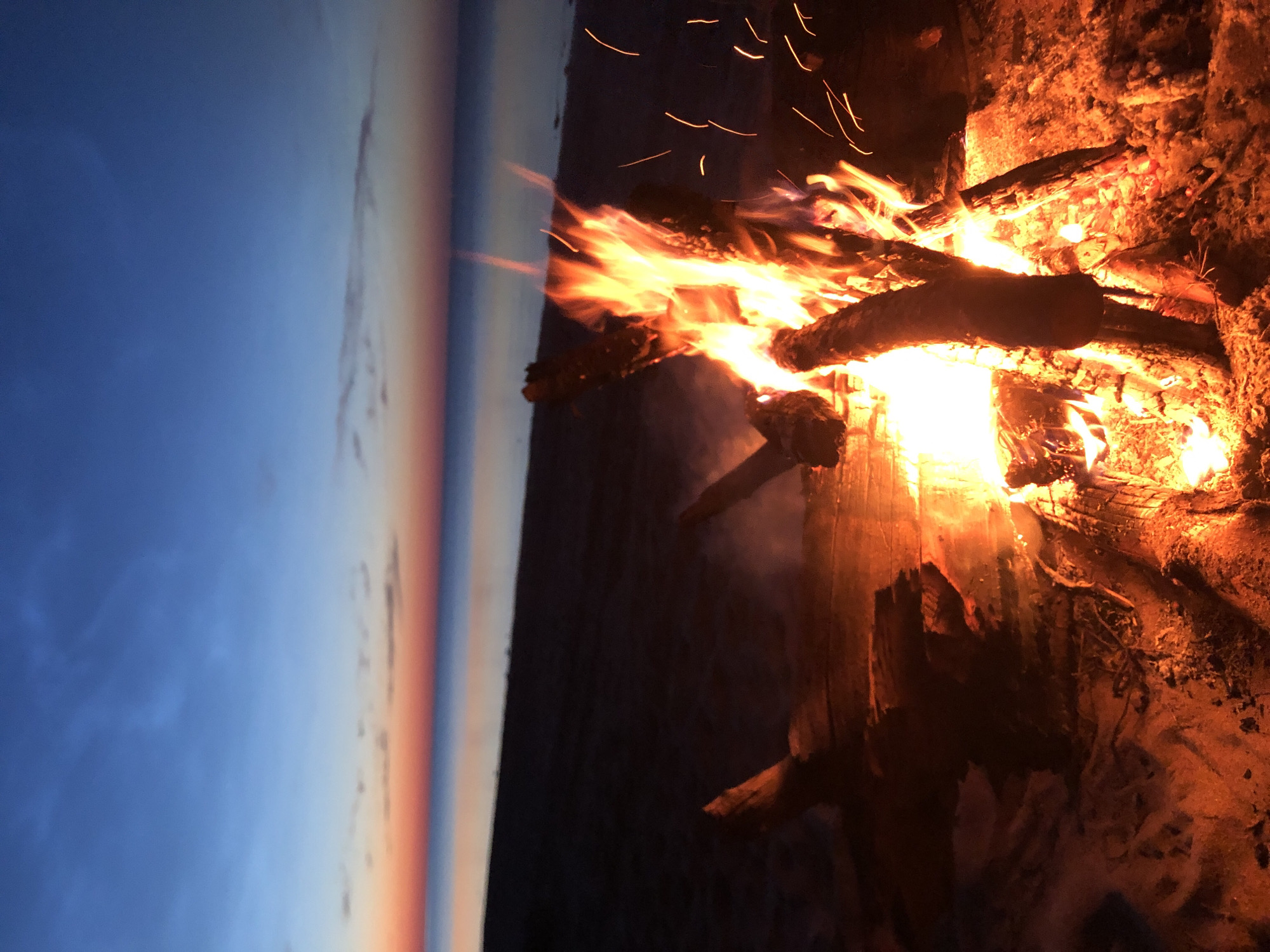 A campfire burns on a sandy beach at dusk. The sky transitions from deep blue at the top to orange and pink at the horizon, meeting the calm, dark water of the sea. The fire is made of logs and is emitting smoke and sparks. Driftwood lies on the sand near the fire. The scene is set in Zaostrov'e, Russia. There are no people in the image.