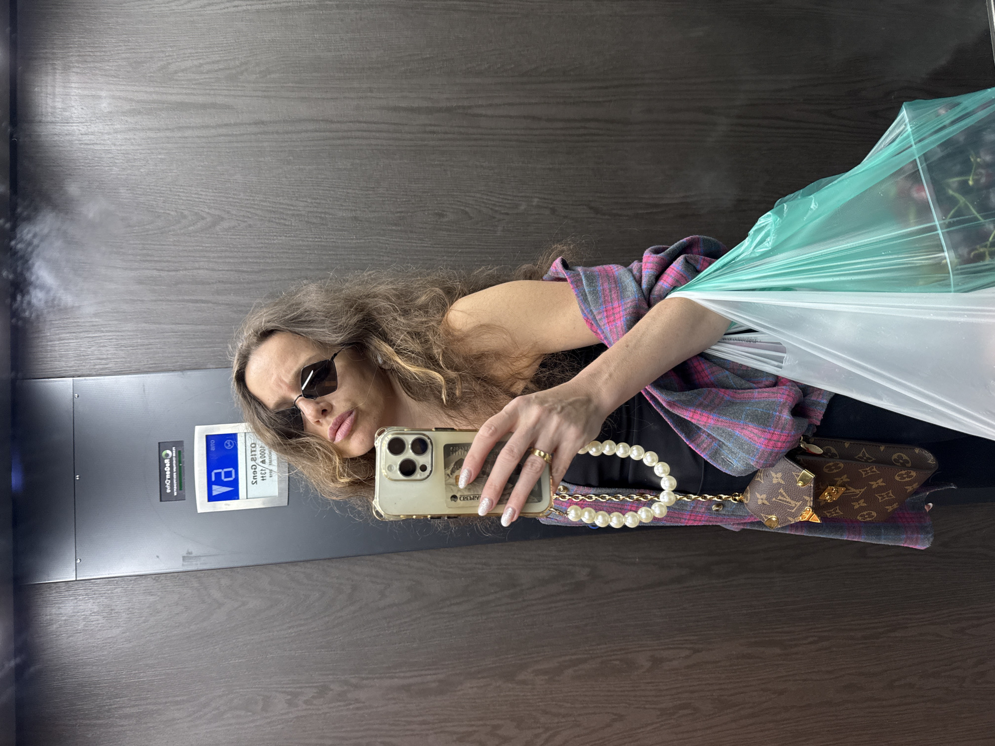 A woman is taking a selfie in an elevator. She has long, wavy brown hair, is wearing sunglasses, and a pink and grey plaid shirt over a black top. She is holding a smartphone in her right hand and a string of pearls dangles from it. A small Louis Vuitton wallet is attached to the pearls. To her left, she is holding a large, clear plastic bag filled with what appear to be grapes. The elevator walls are a dark wood grain, and the elevator door has a silver panel with some signage. One sign is blue with white text that appears to be an elevator button. Another sign is white with black text, possibly instructions or information. The image appears to be taken indoors, likely in a residential building or hotel. The lighting is artificial.