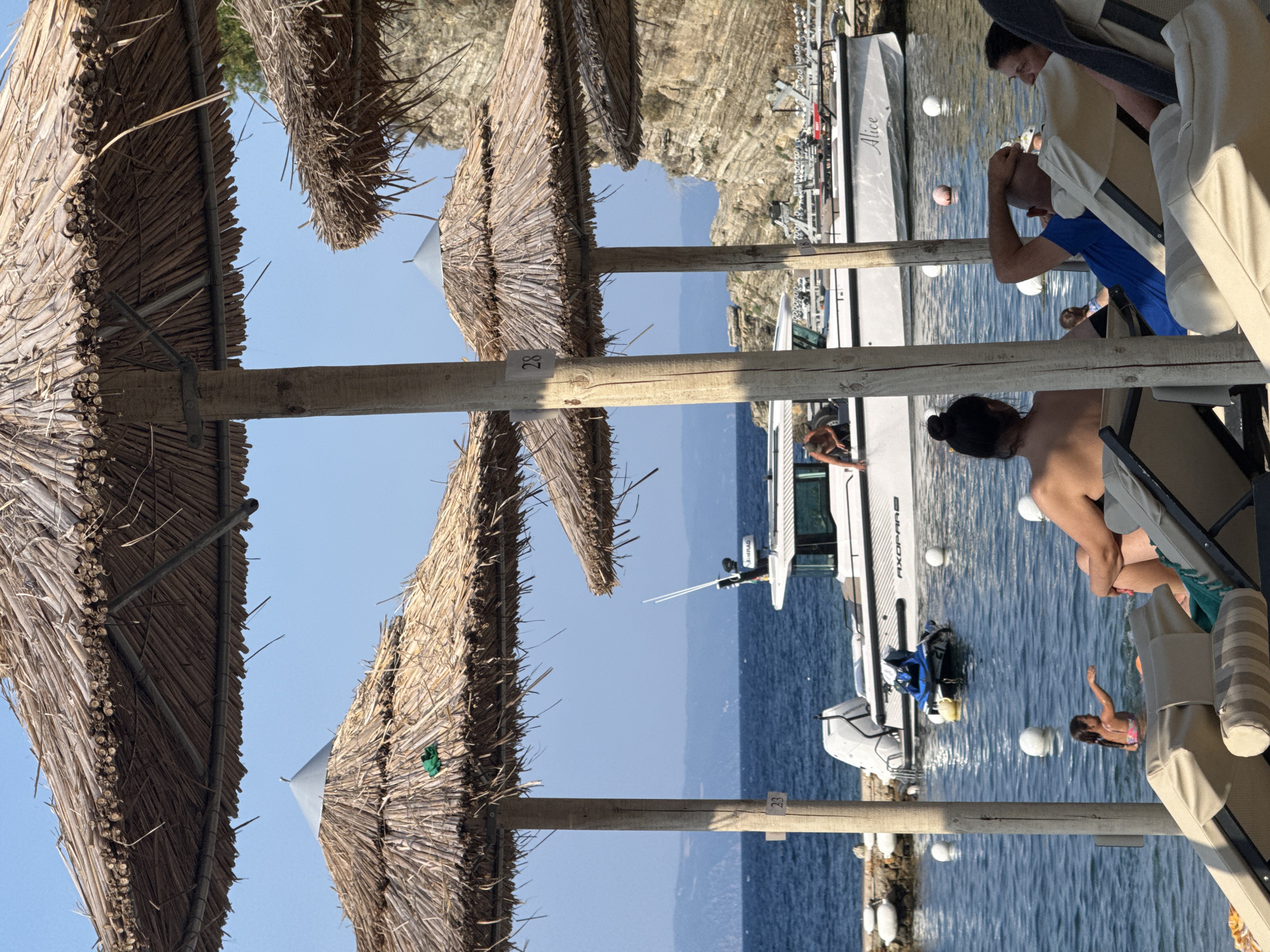 The image depicts a sunny day at a beach in Sithonia Municipal Unit, Greece. In the foreground, several straw umbrellas provide shade for beach chairs where people are relaxing. In the middle ground, the clear blue water of the Aegean Sea is visible, with a boat docked near the shore. A young girl is playing in the water. In the background, rocky cliffs rise from the sea, characteristic of the Sithonia peninsula's coastline. The overall atmosphere is one of leisure and vacation.