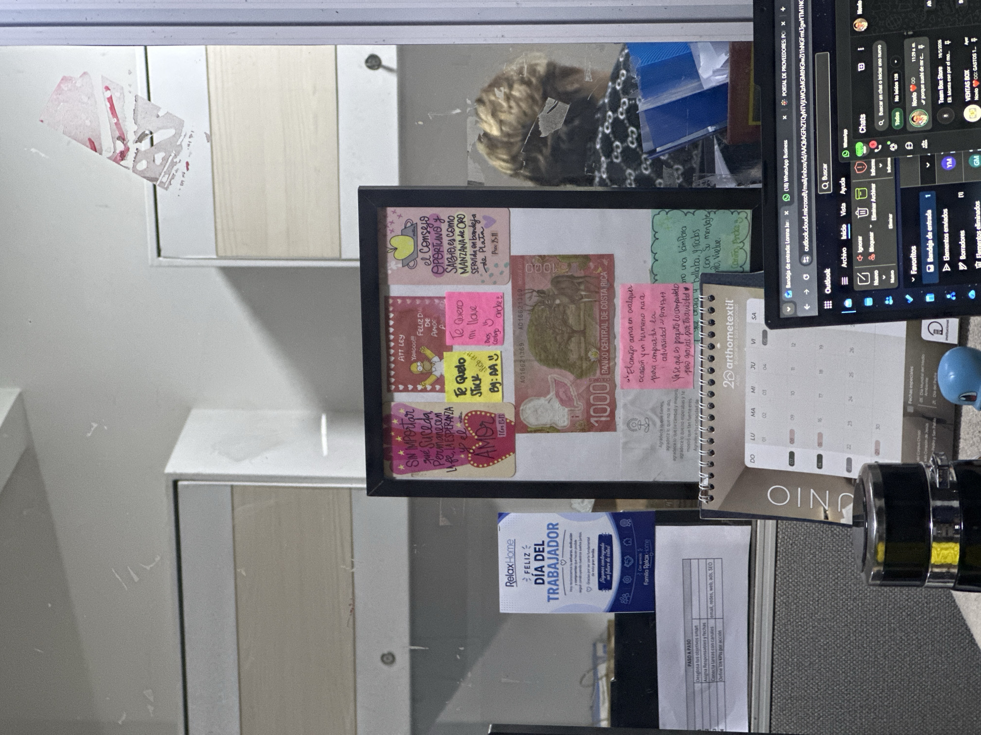 The image shows a desk with a bulletin board, a calendar, and a laptop. The bulletin board has several colorful notes and a Costa Rican 1000 colones banknote. A person with blond hair is partially visible in the background, seated at the desk, looking down at their laptop. The laptop screen displays an email interface and a WhatsApp chat window. The environment appears to be an office. The location is not discernible from the image provided.
