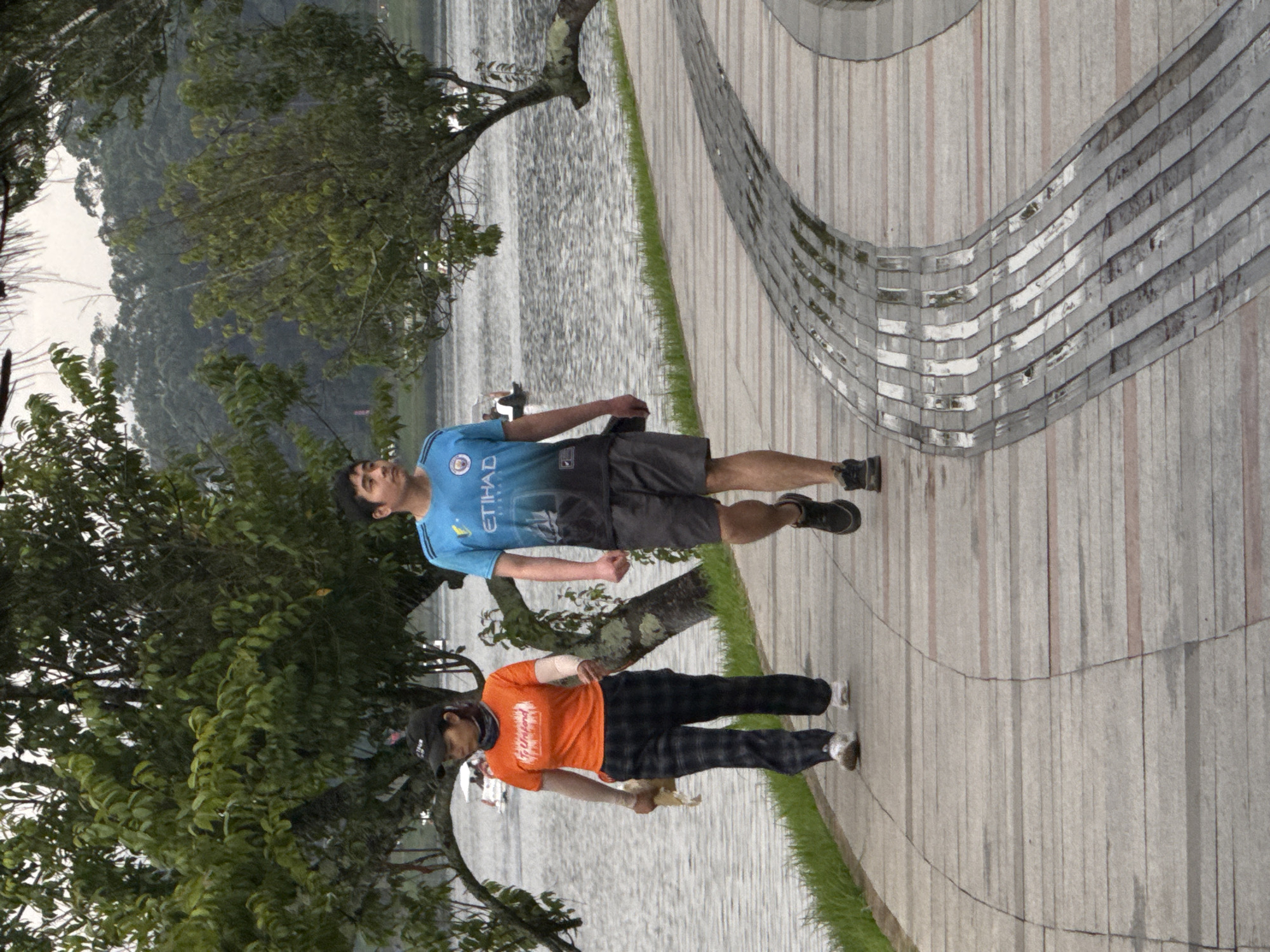 Two individuals are walking on a paved pathway beside a body of water. The person on the left is a woman wearing an orange t-shirt, black and white plaid pants, a black baseball cap, and white sneakers. She is holding a light-colored bag. The person on the right is a man wearing a blue and black t-shirt with a graphic print and black shorts, and black athletic shoes. He is walking slightly ahead of the woman. The pathway is made of light-colored concrete slabs with darker, textured strips. There is a grassy area between the pathway and the water. Trees with green foliage are visible on the left and right sides of the scene, with some branches extending over the pathway. In the background, across the water, is a vegetated hillside. The sky appears overcast. This scene is consistent with pathways around Xuan Huong Lake in Da Lat, Vietnam, a popular public recreational area.