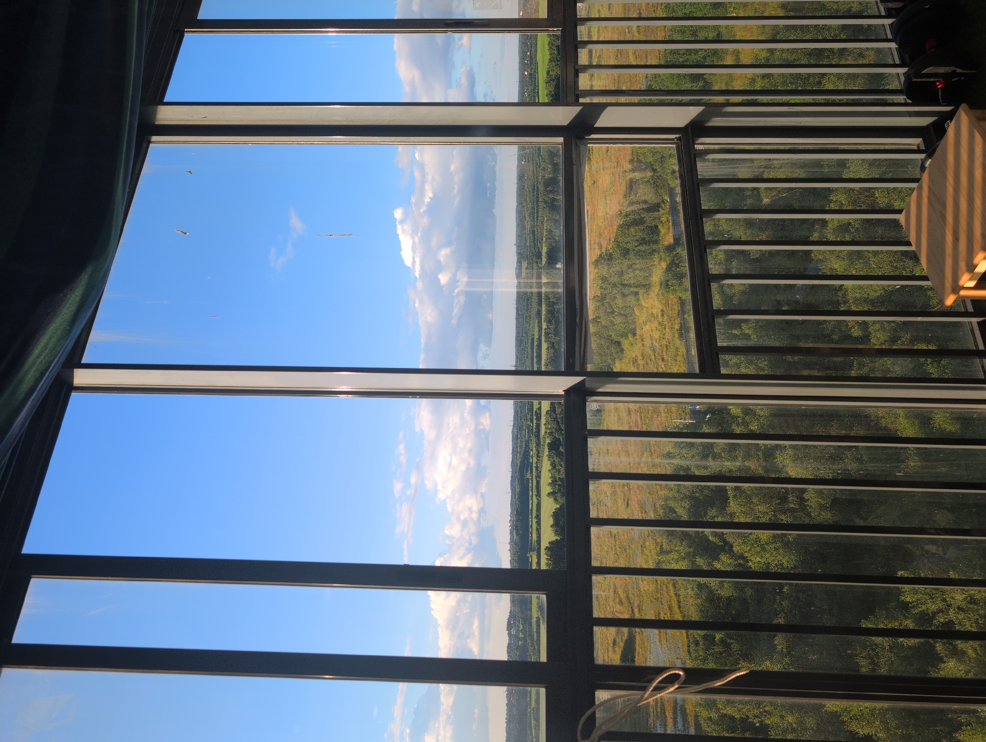An interior view looking out of a building with large windows onto a rural landscape. The foreground features a metal railing and a portion of a wooden table, with some dark objects, possibly bags, to the right. The middle ground shows a grassy field with scattered trees and some bodies of water. The background consists of rolling hills covered in trees and fields, with a few buildings and power lines visible in the distance under a bright blue sky with scattered white clouds. The scene appears to be located in the vicinity of Bugry, Russia, given the architectural style of the windows and the nature of the landscape.