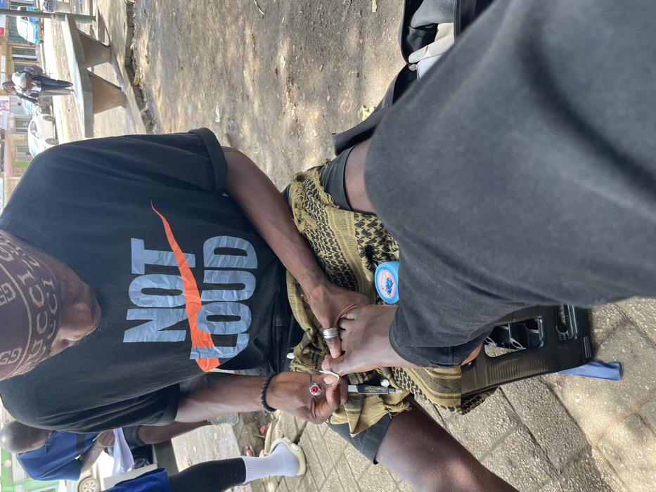This media shows an outdoor scene in Luanda, Angola, during the daytime. The central focus is on two individuals engaged in a pedicure service on a paved surface, likely a sidewalk or informal market area.
The person performing the pedicure is seated, wearing a black t-shirt with the text "NOT GOOD" prominently displayed, where "GOOD" is crossed out by a large orange checkmark resembling a stylized swoosh. They also wear a patterned bandana or headwrap featuring the "GUCCI" logo. Their left arm, with visible veins, rests on a patterned scarf draped over their lap. They are holding nail clippers and are actively working on the foot of the second individual. A ring is visible on their right hand and a beaded bracelet on their wrist.
The second individual, whose legs are visible, is receiving the pedicure. They are wearing dark pants, and their left foot rests on a black plastic stool or stand. A small blue container is placed near their foot on the patterned scarf.
In the background, other people are partially visible, including one person in a blue shirt and white socks. Cars and what appears to be part of a bench or structure can also be seen further down the street, suggesting an urban or semi-urban environment. The bright natural light indicates it is daytime, with clear visibility and no signs of adverse weather. The overall impression is an informal street-side service.