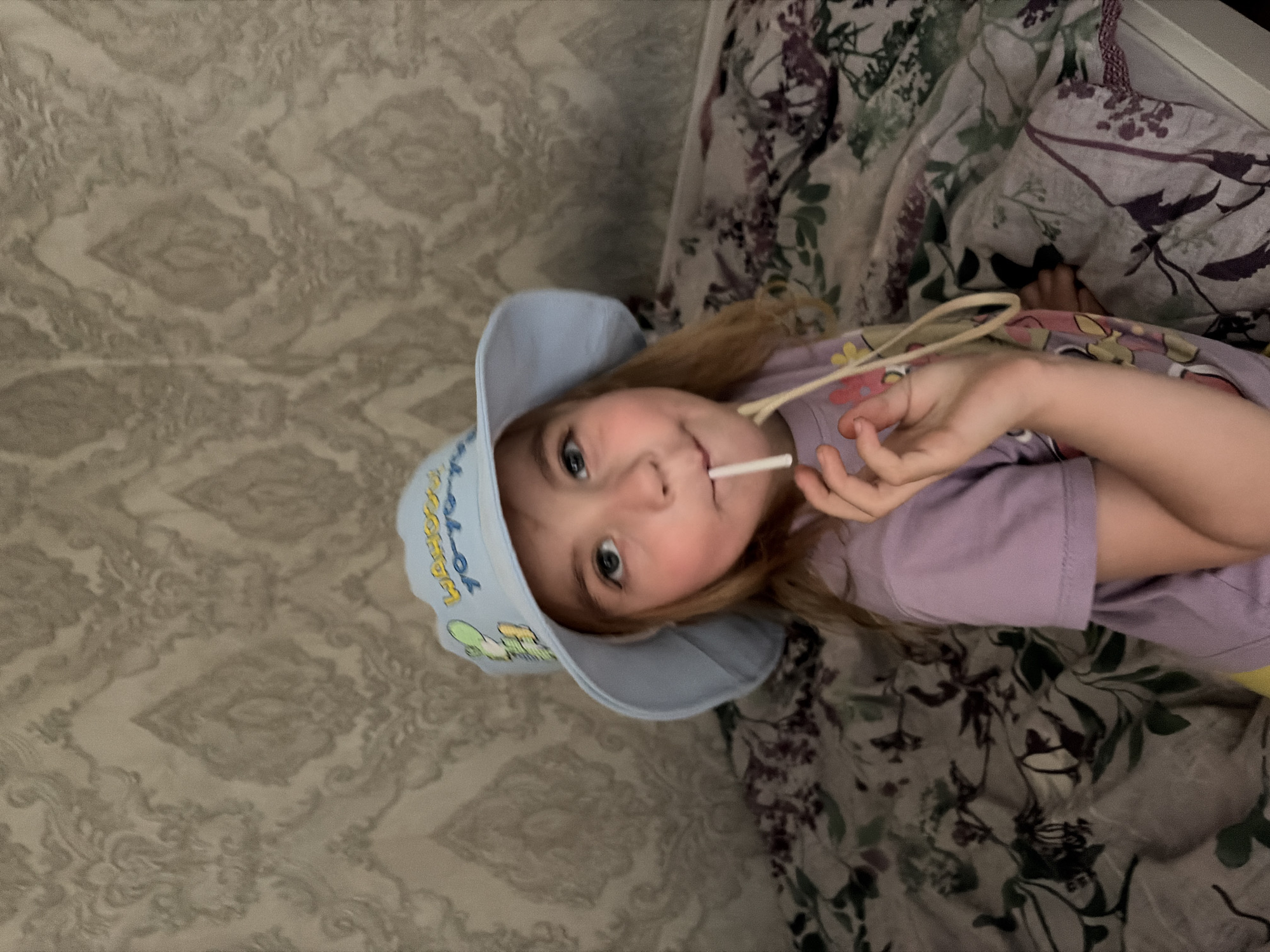 A young girl with blonde hair and blue eyes, wearing a light blue bucket hat and a purple t-shirt, is sitting on a bed. She is holding a lollipop to her mouth. The background shows a patterned wallpaper and a floral comforter on the bed. The scene appears to be indoors, likely a bedroom. There is no information in the image to suggest a location in Tambov, Russia.