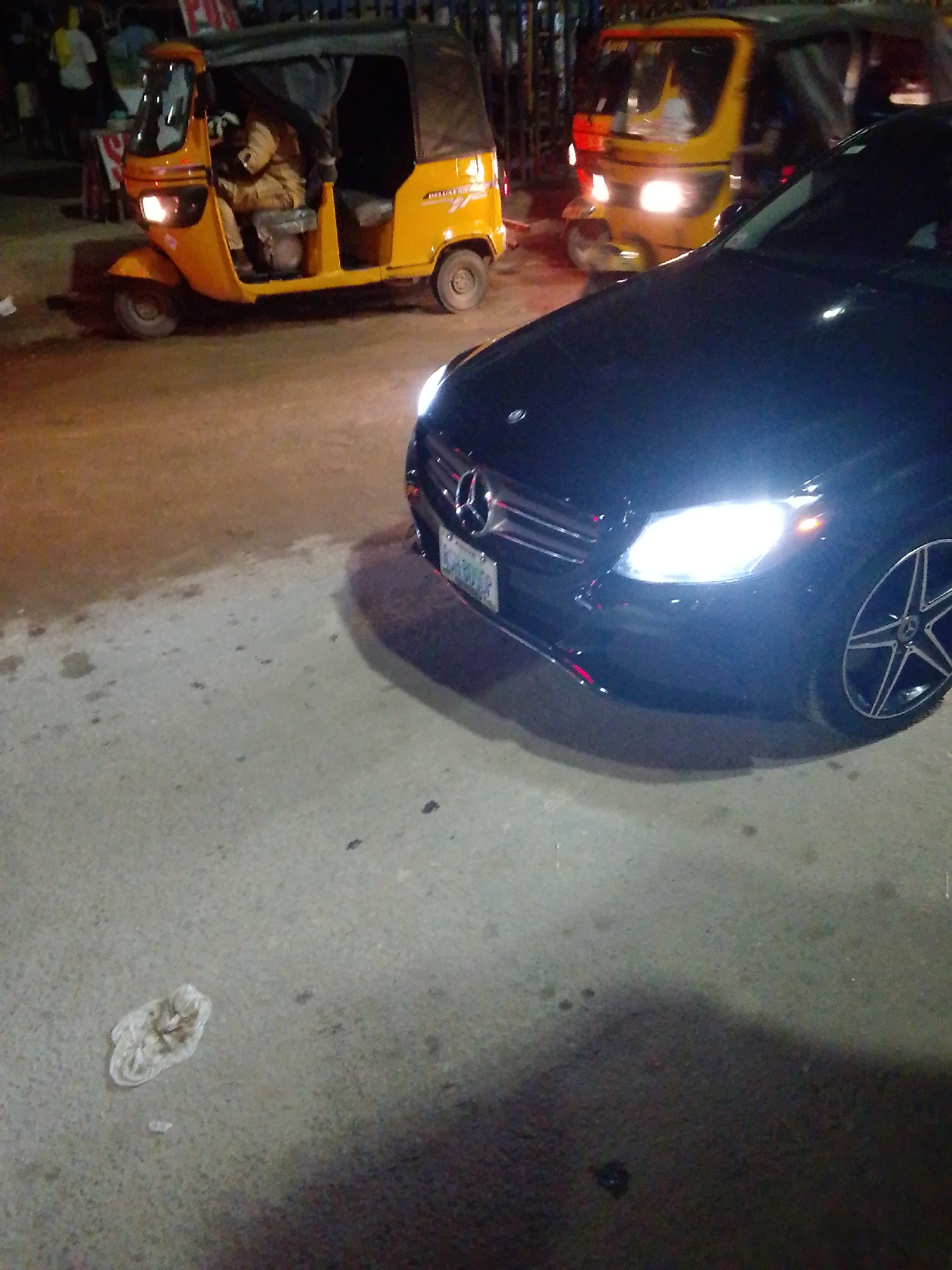 A nighttime scene in Lagos, Nigeria, showcases a black Mercedes-Benz C-Class parked on a concrete surface. The car's headlights are illuminated, and its front is angled toward the viewer. Behind the Mercedes, two yellow tricycles (also known as "keke napep" or "tuk-tuks") are visible, their headlights also on. The tricycle on the left has a driver inside. In the background, indistinct figures suggest a bustling street scene, likely indicating a commercial area. Litter is visible on the concrete surface near the car.