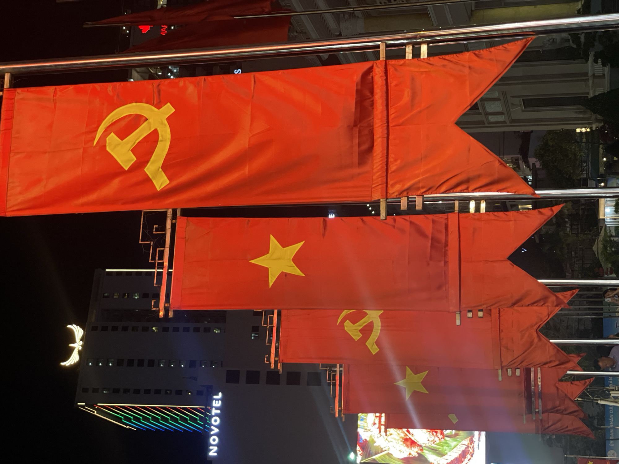 The image depicts a nighttime urban scene in Nha Trang, Vietnam, characterized by a prominent display of red flags. Multiple flags are arranged vertically, attached to horizontal metal railings, and appear to be hanging as part of a public decoration or celebration.
Several flags feature the yellow hammer and sickle emblem, a symbol of communism. Interspersed among these are flags displaying a large yellow five-pointed star on a red background, which is the national flag of Vietnam. The flags are distinctively shaped with a swallowtail or pennant at their flying end.
In the background, a tall building is illuminated against the dark sky. The word "NOVOTEL" is visible in vertical, white neon letters on its side. Above this, a large, glowing, white bird-like emblem is affixed to the building's facade. The lower part of the Novotel building features horizontal strips of colorful lighting. A partial sign displaying the Vietnamese text "ỦY BAN NHÂN DÂN" (People's Committee) is also discernible in the lower right, suggesting a government presence nearby. The overall setting is brightly lit, indicative of a busy city area during the evening, likely adorned for a national or commemorative event.