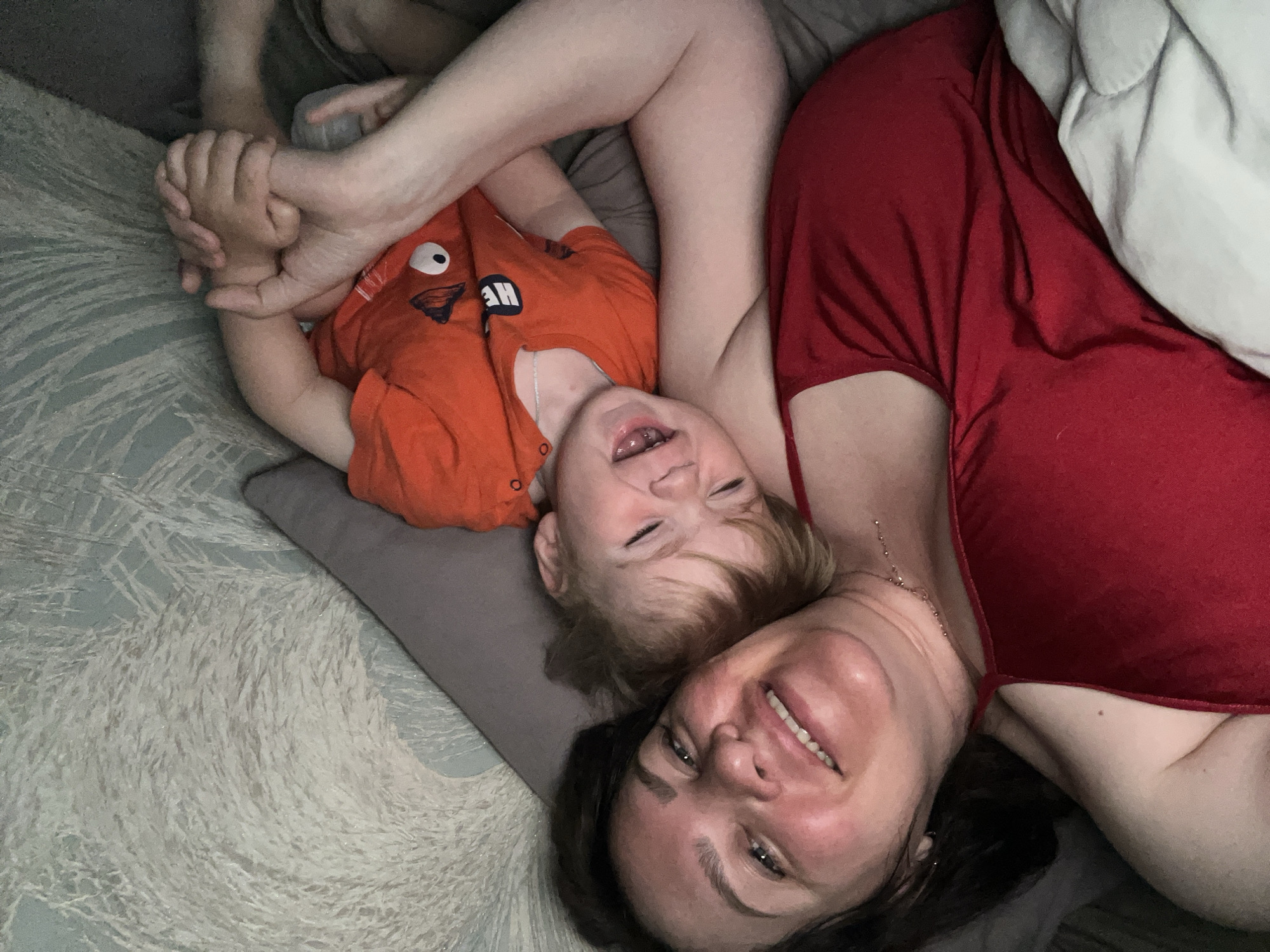 In Noviy, Russia, an overhead shot captures a mother and child in bed. The mother, with brown hair and fair skin, wears a red camisole and a necklace. She is smiling and her arm is around the child. The child, with light brown hair, wears an orange t-shirt and is leaning against the mother, with eyes closed and mouth open in a smile. They are lying on a gray pillow, with a white blanket visible to the left and a textured light blue wall to the right. The overall scene conveys a moment of intimacy and affection between mother and child.