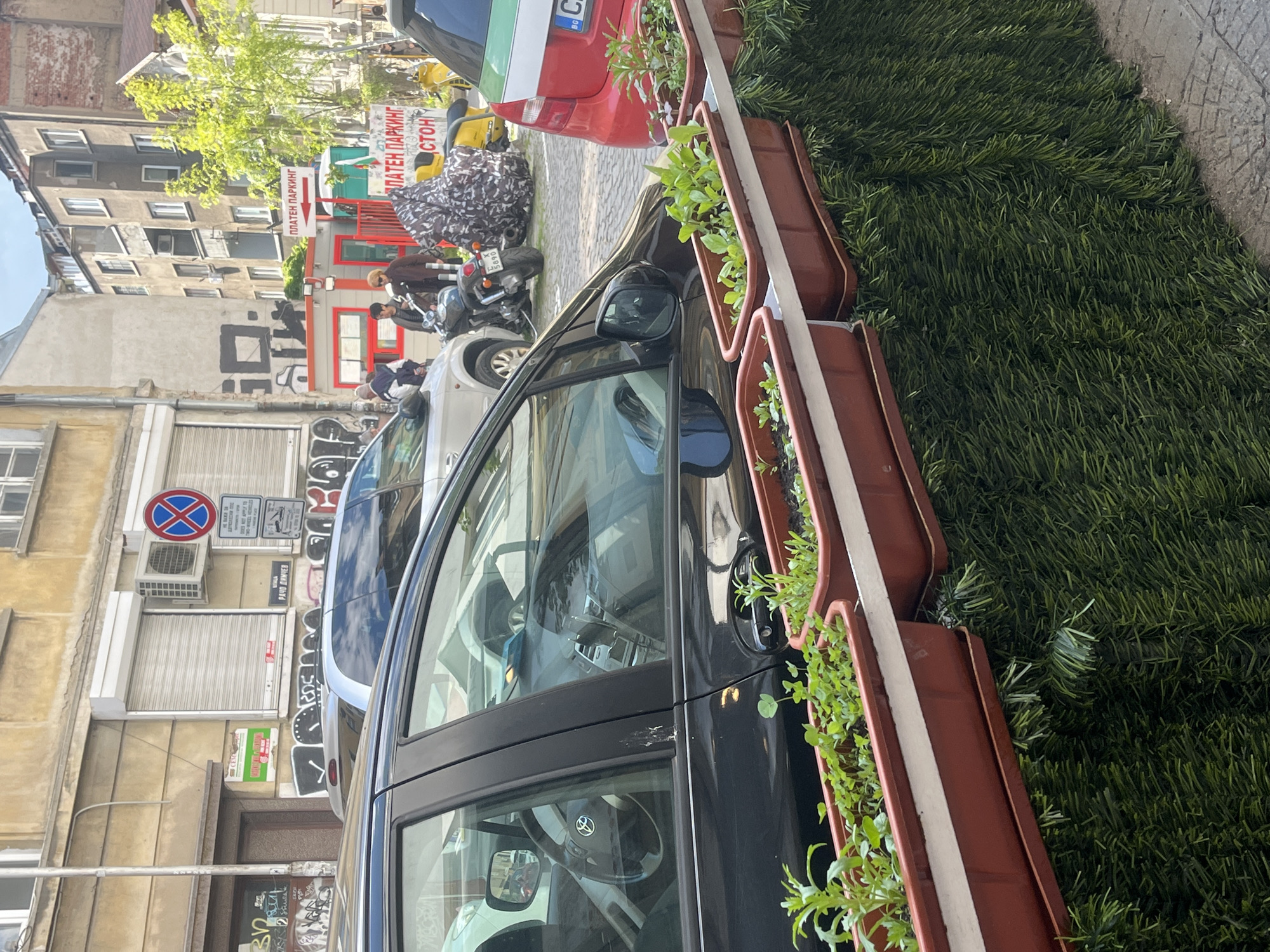The image depicts a street scene in Sofia, Bulgaria, with a black car parked in the foreground, adorned with flower boxes on its side. The boxes are filled with green plants. In the background, there are other parked vehicles, including a red car and motorcycles, near a red and white building with signs in Bulgarian Cyrillic script. The street is lined with buildings that are several stories high. A street sign indicating "ПЛАТЕН ПАРКИНГ" (Paid Parking) is also visible. Artificial green turf covers the area beside the flower boxes. There is a parking prohibition sign visible on the wall of a building.