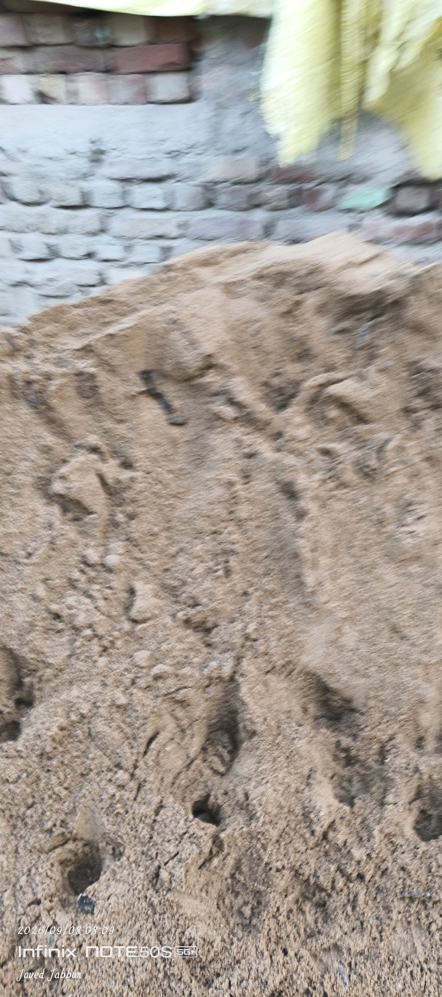 A pile of sand is in the foreground of the image, with a blurred brick wall and yellow material in the background. The sand appears to be disturbed, with footprints and indentations visible. A dark object resembling a nail or small piece of metal is embedded in the sand. In the lower left corner of the image, there is a timestamp and text that reads "2026/09/08 08:09 Infinix NOTE30SG+| Javed Jabbar". The overall impression is of a construction or outdoor area in Bahera, India, with construction materials and signs of activity.