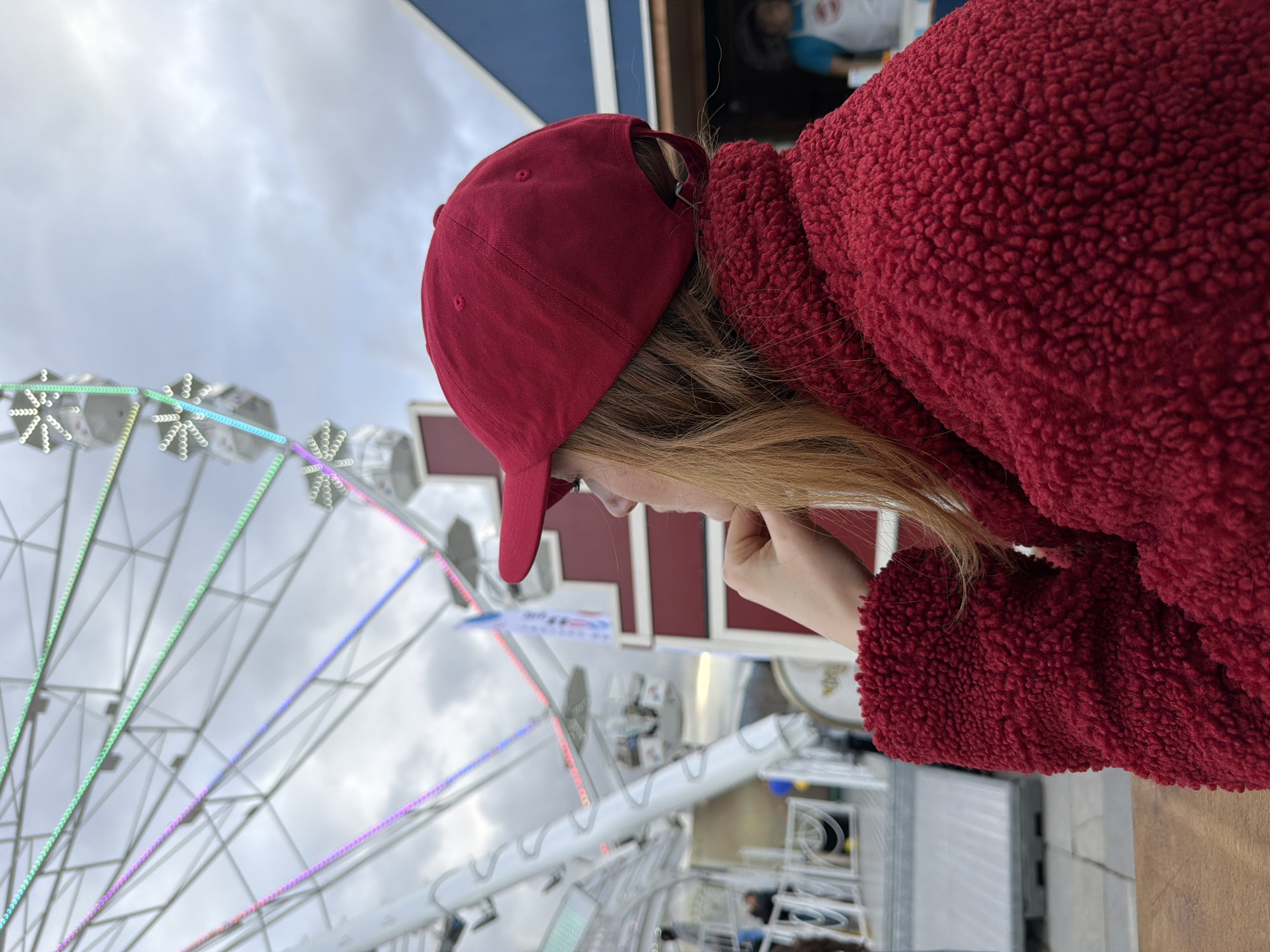 A woman wearing a red fleece jacket and a red baseball cap stands sideways, looking at a ferris wheel. The ferris wheel is white and decorated with colorful lights. The scene is likely located in Sofia, Bulgaria, as this is the location specified in the prompt. The woman appears thoughtful, resting her chin on her hand.