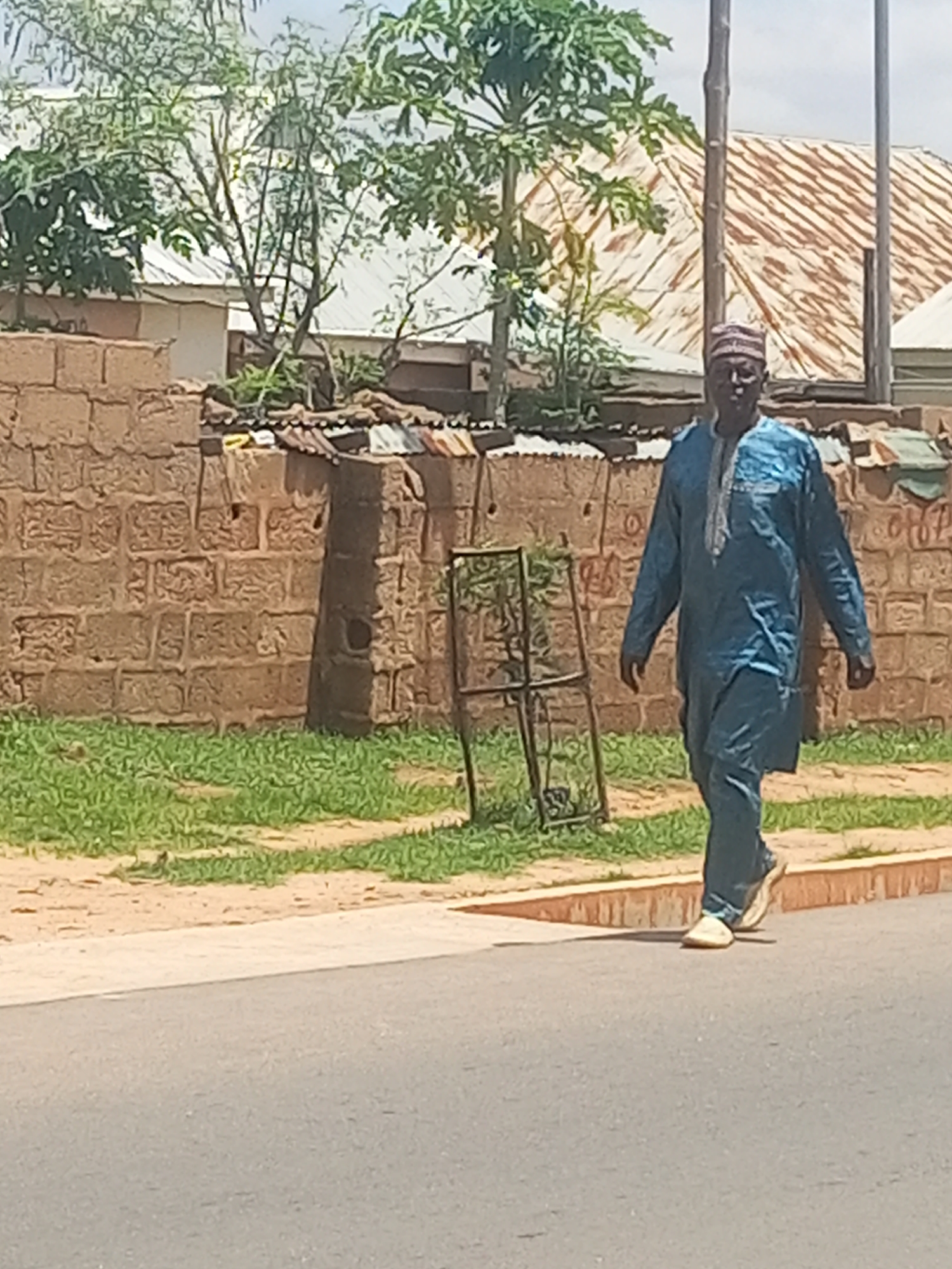 A man in a blue traditional outfit and a cap walks on a paved road in front of a brick wall and some green grass. Behind the wall, a building with a corrugated metal roof and some trees are visible. The scene appears to be in a rural or semi-urban area, possibly Tempure, Nigeria, judging by the style of dress and architecture. The man is walking towards the right of the frame.