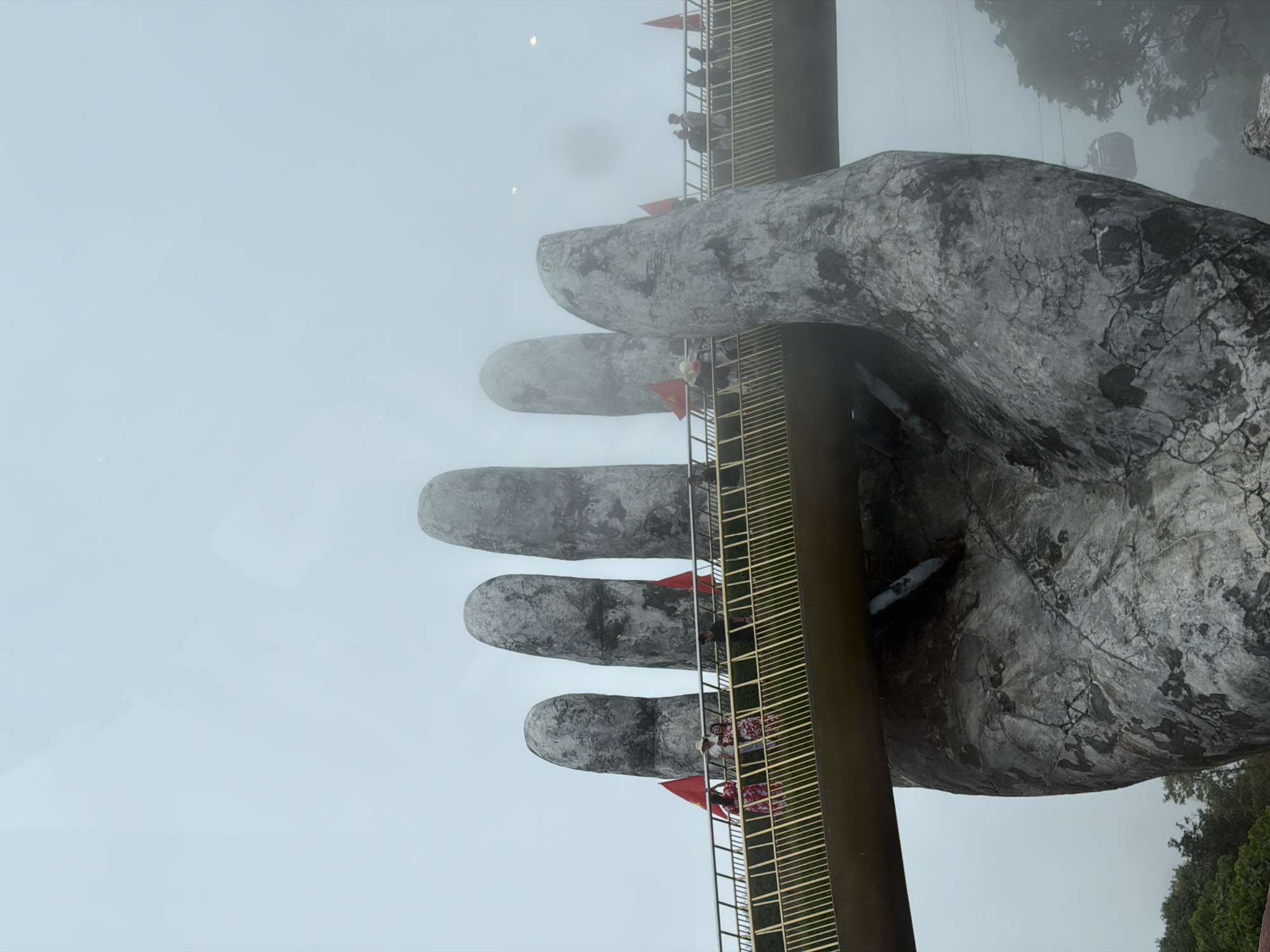 The image depicts the Golden Bridge in Đà Nẵng, Vietnam, held by two giant stone hands. A walkway with metal railings spans between the hands, with people walking along it. The bridge and hands are partially obscured by fog. Vietnamese flags are mounted on the railings. Foliage is visible in the background. The sky is overcast.