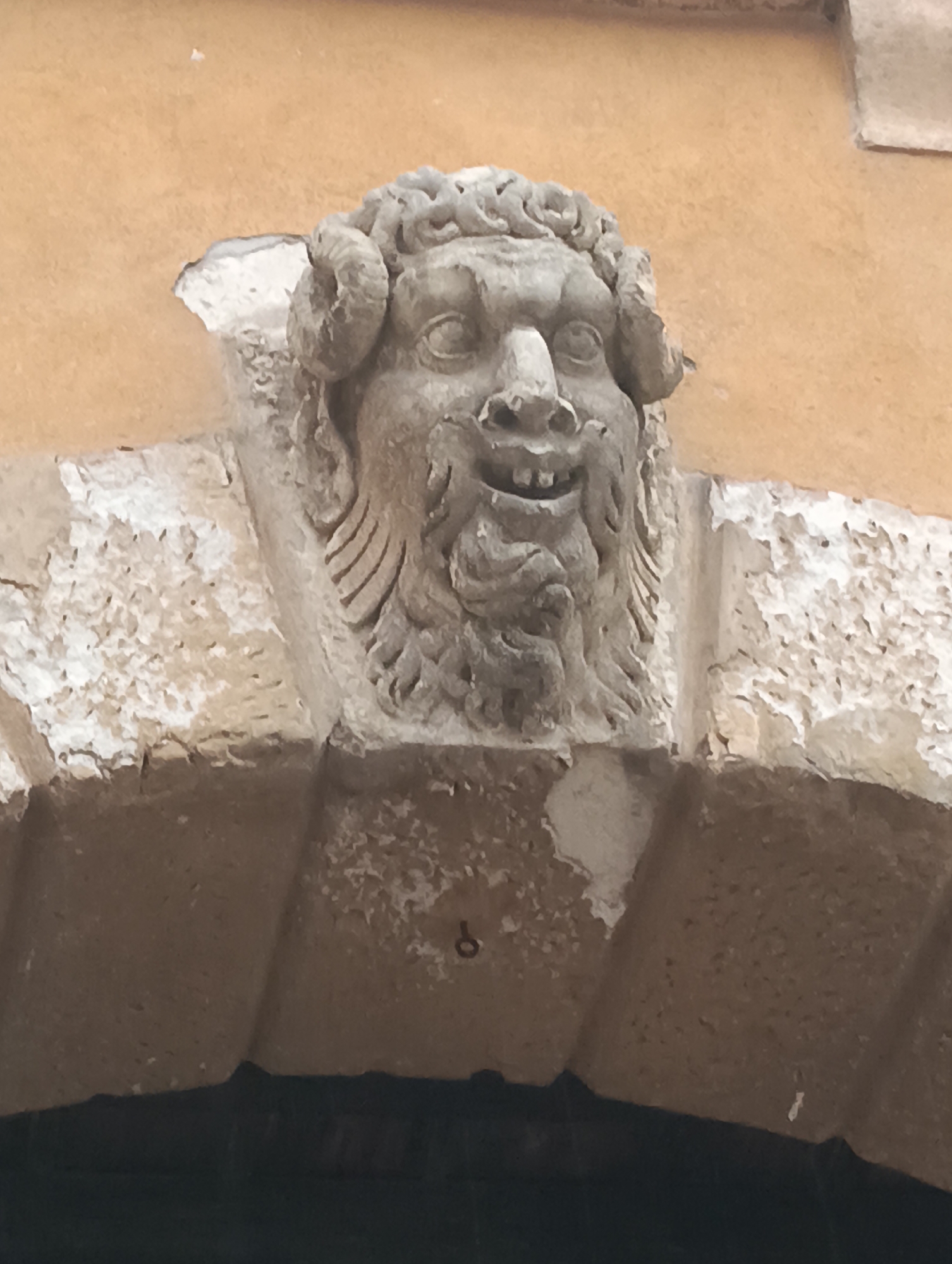 The image shows a weathered stone carving of a grotesque face with horns, a beard, and a gaping mouth with visible teeth, affixed to the facade of a building. The carving is situated above an arched opening. The building's wall is a rough, orange-toned stucco. The stone carving exhibits signs of age and erosion, with chipped edges and a rough texture. The architectural style suggests it is an ancient building, possibly in an historical district. This carving is located in Verona, Italy, specifically at 43 Via Mazzini, the former location of the Antica Trattoria della Scala.