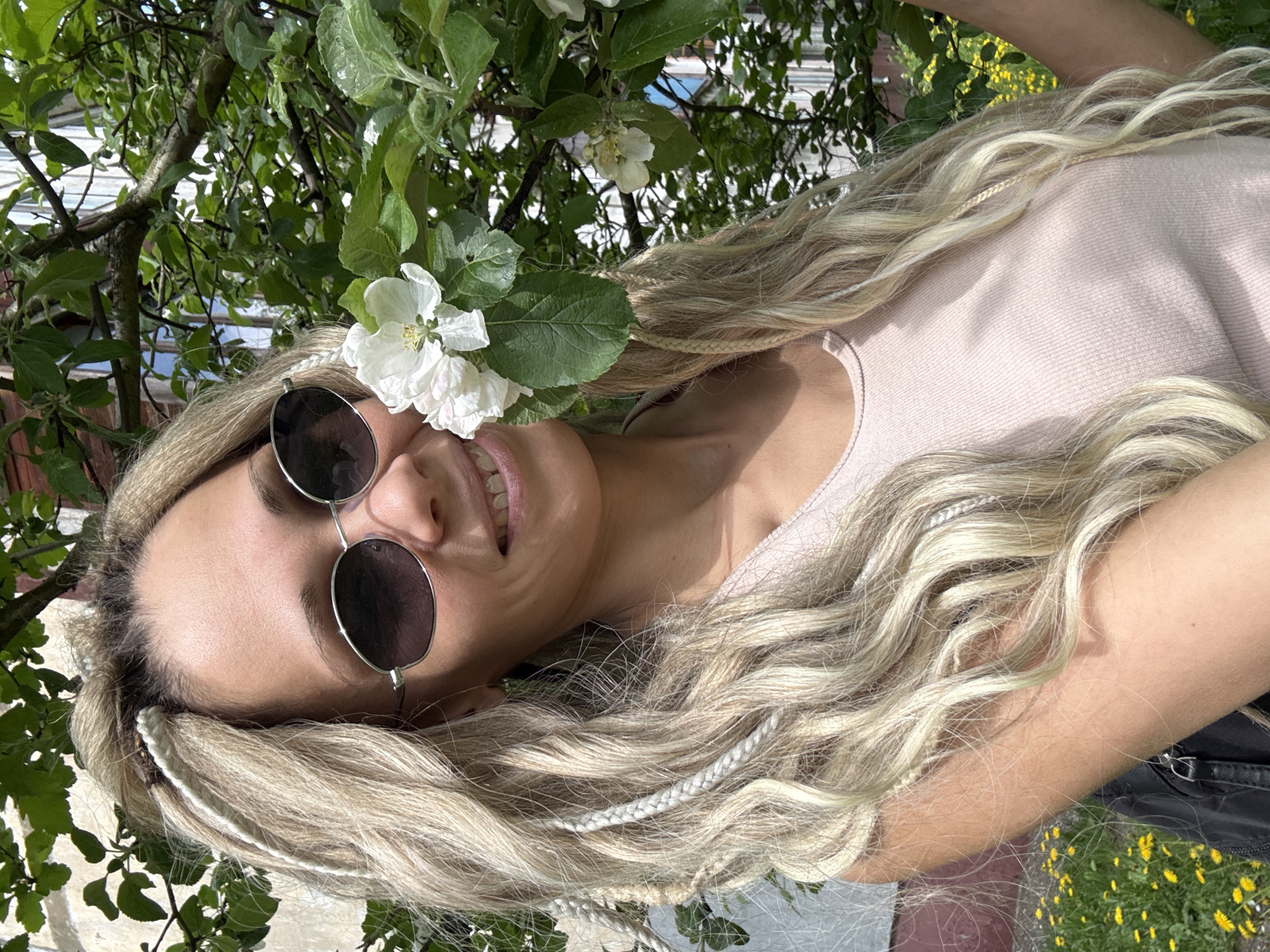 A light-skinned woman with blonde hair and small white braids is smiling in the image. She is wearing round sunglasses with silver frames. A flowering tree branch, with one white flower prominently displayed, is positioned near her face. She wears a light pink top. Dandelions are visible in the background, suggesting an outdoor location. The image is rotated.