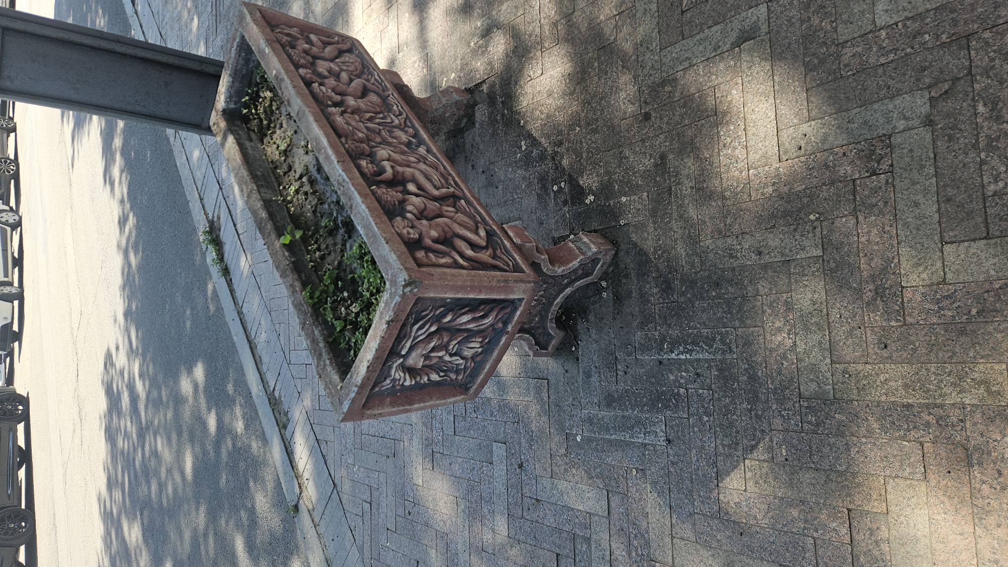 An outdoor, high-angle shot depicts a large, ornate rectangular planter situated beside a city street. The planter, made of a reddish-brown material, features intricate carvings on its exterior, potentially depicting human figures or foliage. Inside the planter, small green plants are visible. To the left of the planter is a metal pole. The planter sits on a brick-paved surface that extends to the right. In the background, a city street and parked cars can be seen under dappled sunlight. This is located in Chișinău, Moldova.