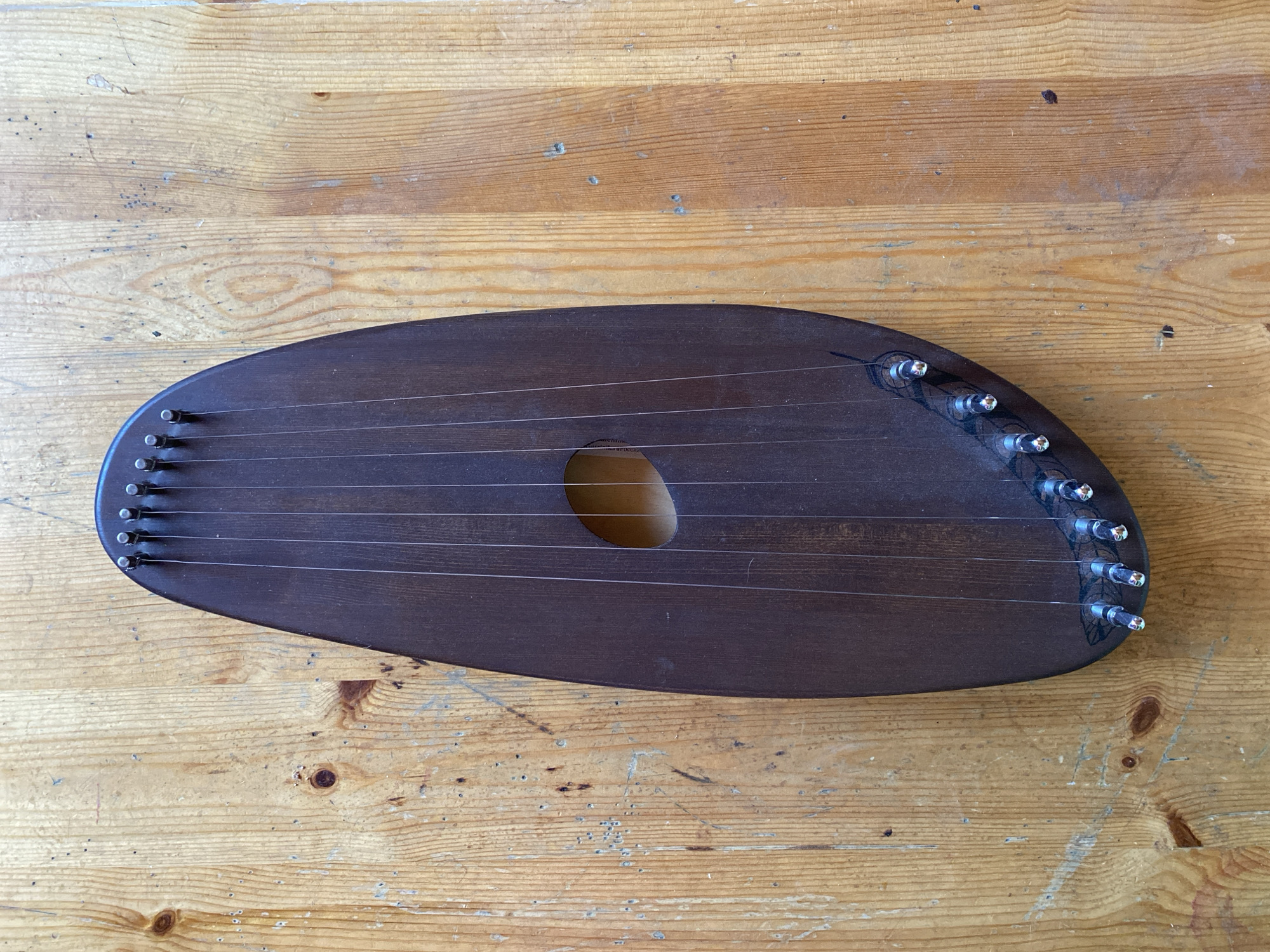 The image shows a top-down view of a dark wooden stringed instrument, possibly a kantele or a lyre, resting on a rustic wooden table. The instrument has an oval shape with a cutout in the center. It is strung with multiple thin strings that run vertically from tuning pegs at the top to tension pins at the bottom. The wood of the table has a natural grain pattern and appears worn, with visible knots and small dark specks. The lighting suggests an indoor setting with natural light source from above. The specific location of Zelenodol'sk, Russia is not ascertainable from the visual information provided in the image. There are no people, actions, or interactions depicted.