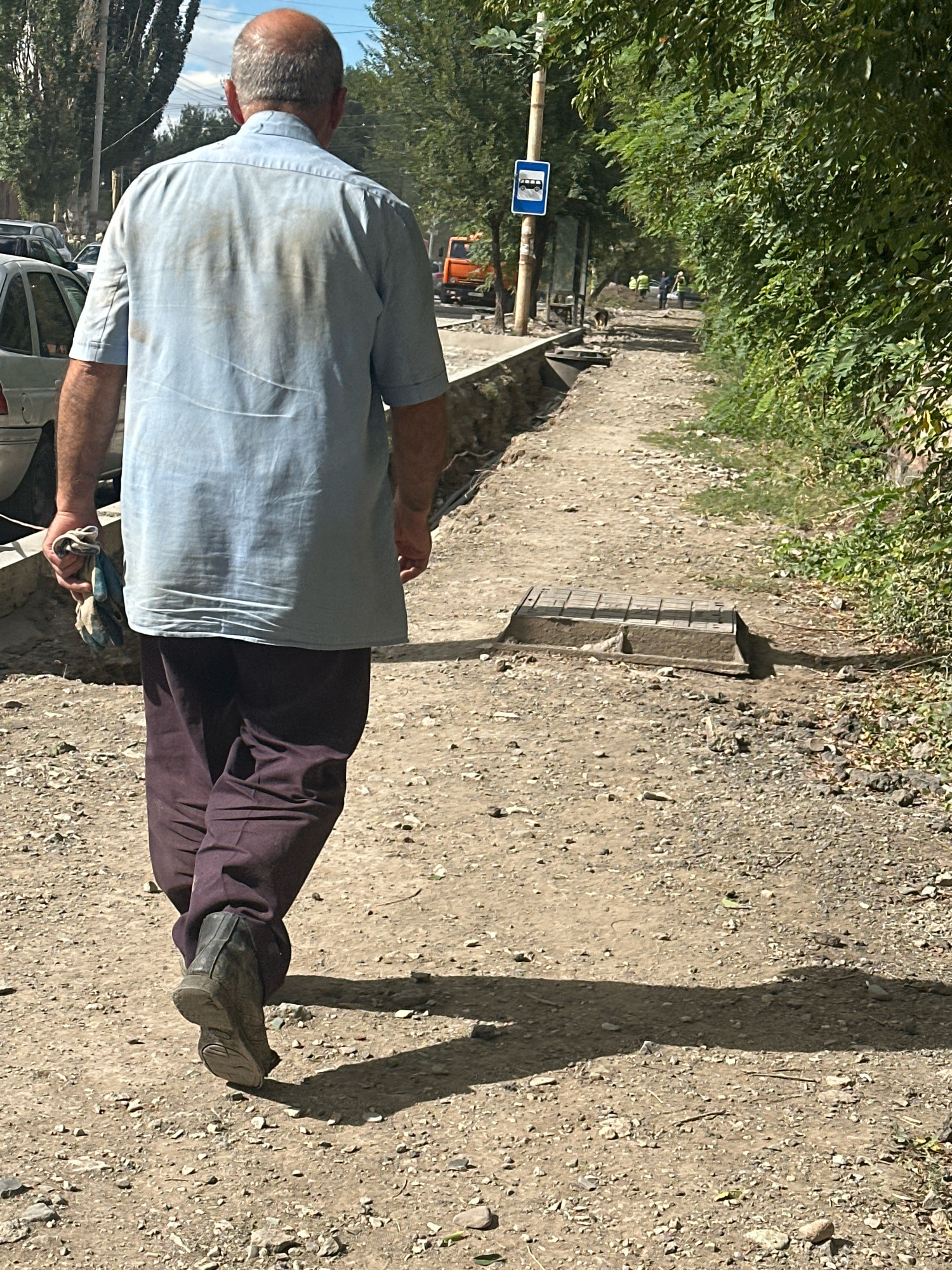 A man, viewed from behind, walks on a dirt path in Gyumri, Armenia. He is bald and wearing a light blue, short-sleeved button-down shirt with dark pants and black boots. He is holding a bundle of cloths in his right hand. The path is unpaved and rocky, with a trench or construction area to his left. A bus stop sign is visible on a pole to the left, with an orange construction vehicle and other workers in the background. Lush green trees line the right side of the path. The scene appears to be during daylight, casting a distinct shadow of the man on the ground.
