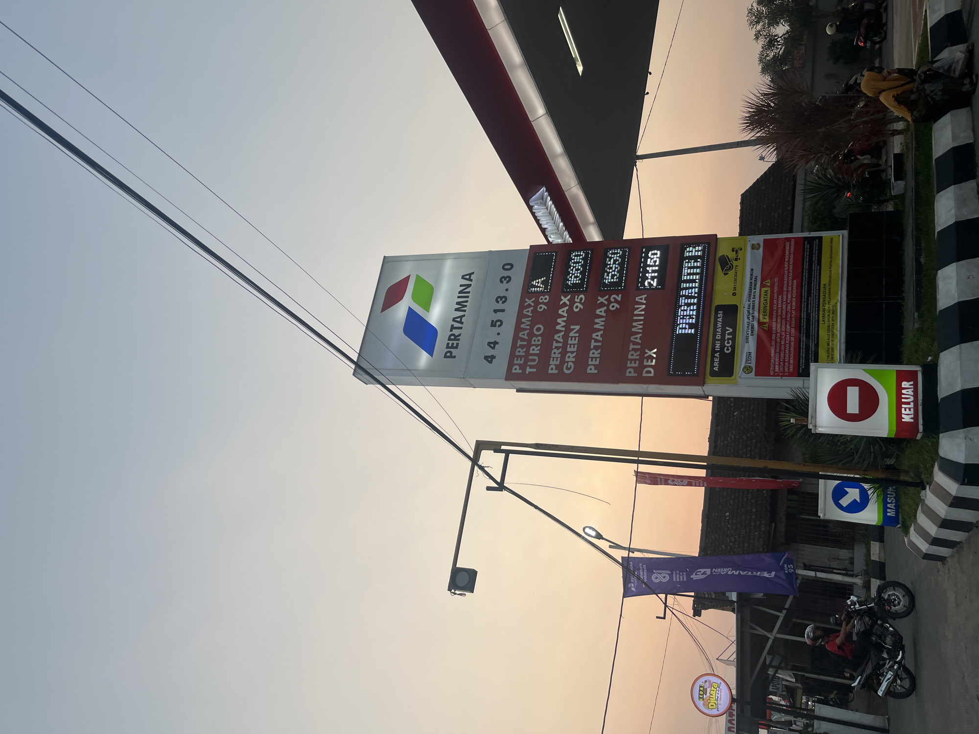 This is a photograph taken at a Pertamina gas station in Boja, Indonesia, during what appears to be sunset or dawn, given the warm, orange glow in the sky. The central focus is a tall, illuminated price board displaying fuel prices for various Pertamina products like Pertamax Turbo, Pertamax Green, and Pertamina Dex, with prices listed in Indonesian Rupiah. The Pertamina logo is prominent at the top of the board.
To the left of the price board, a street light with a speaker hangs over the road. Two colorful banners, one purple and one red, are also displayed, with the purple banner featuring the Pertamina logo and text related to "Pertamina Green Ron 95."
In the foreground, a man on a motorcycle is riding away from the camera. The road in front of the gas station is marked with black and white striped curbs and traffic signs indicating "Masuk" (Entrance) with a blue arrow pointing right and "Keluar" (Exit) with a red circle and a white horizontal bar.
Behind the price board and banners, a building with a dark, tiled roof is visible, along with some vegetation. A few other people are seated on the curb on the right side of the image. The overall scene depicts a typical day at an Indonesian gas station, with vehicles, signage, and the ambient light of the sky setting the mood.