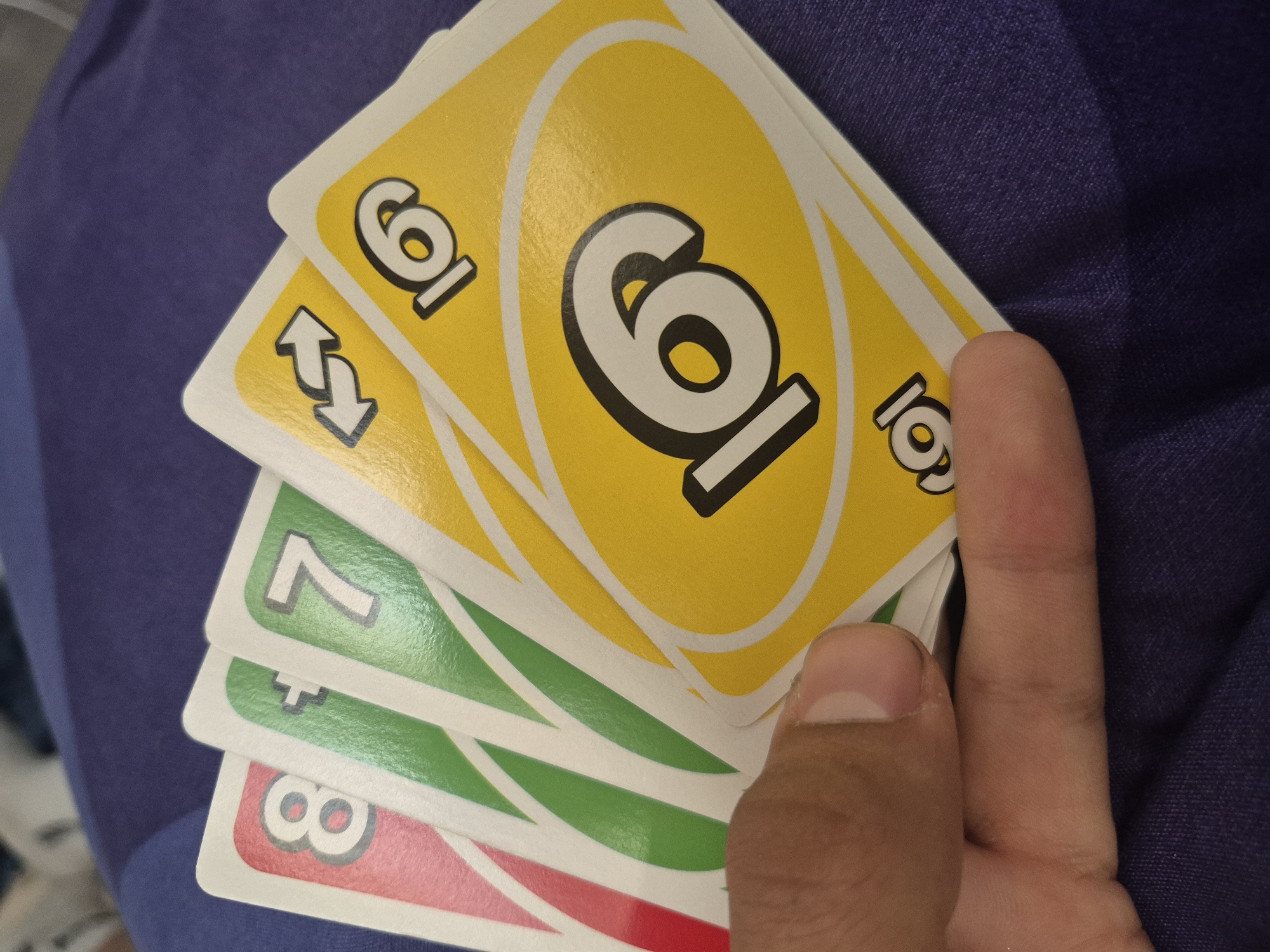 A hand holds five Uno playing cards against a textured blue background. The top card is yellow with a "6". Underneath is a yellow reverse card and green cards labeled "7" and "8", as well as a 5th card. Location: Unknown, Unknown.