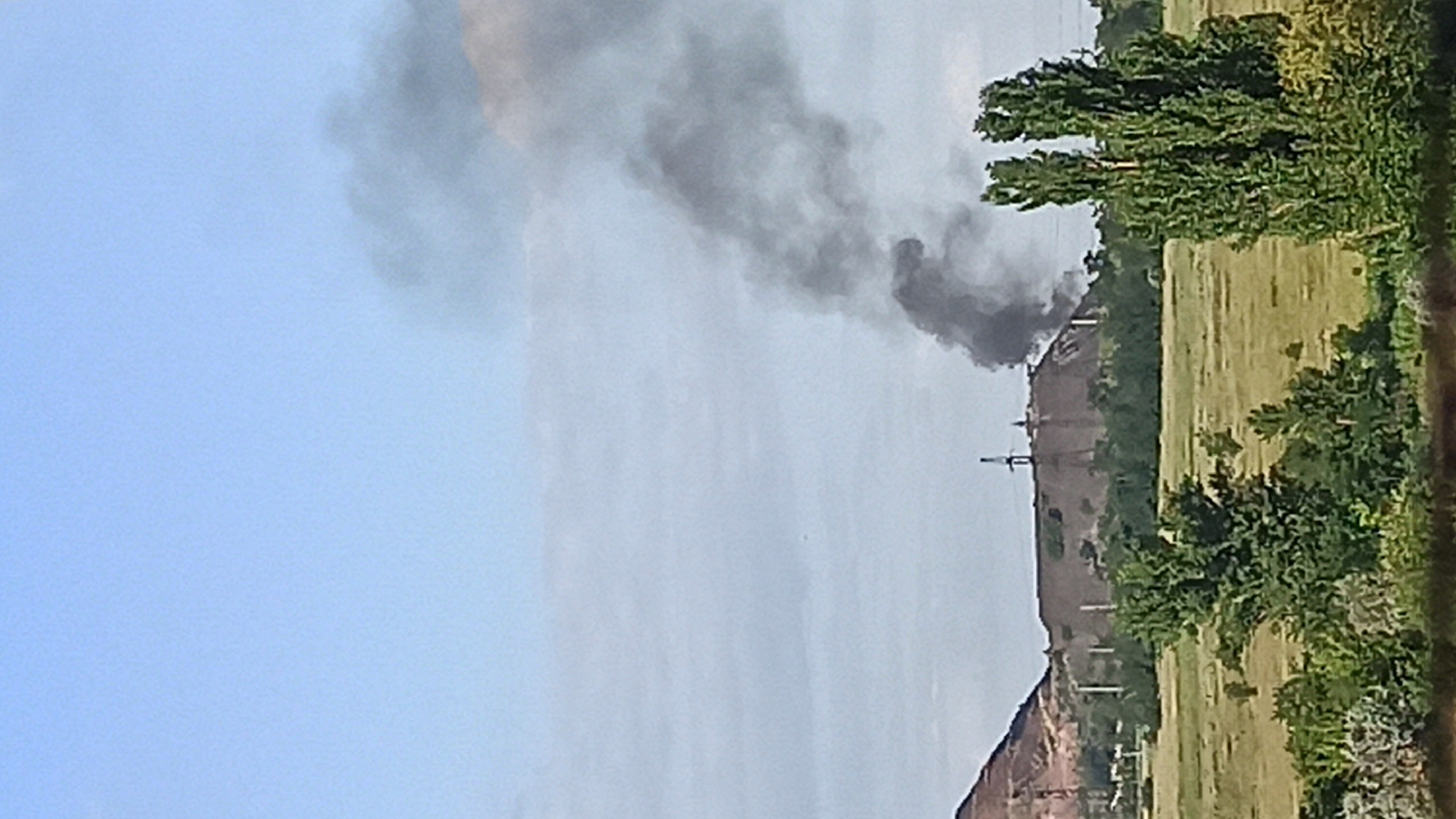In a distant, open field under a hazy blue sky, a large slag heap or industrial waste pile is visible. Smoke billows from the top of the pile, obscuring a portion of the sky. In the foreground, lush green trees and foliage line the bottom of the frame. The scene appears to be an industrial area or a location where industrial waste has accumulated.