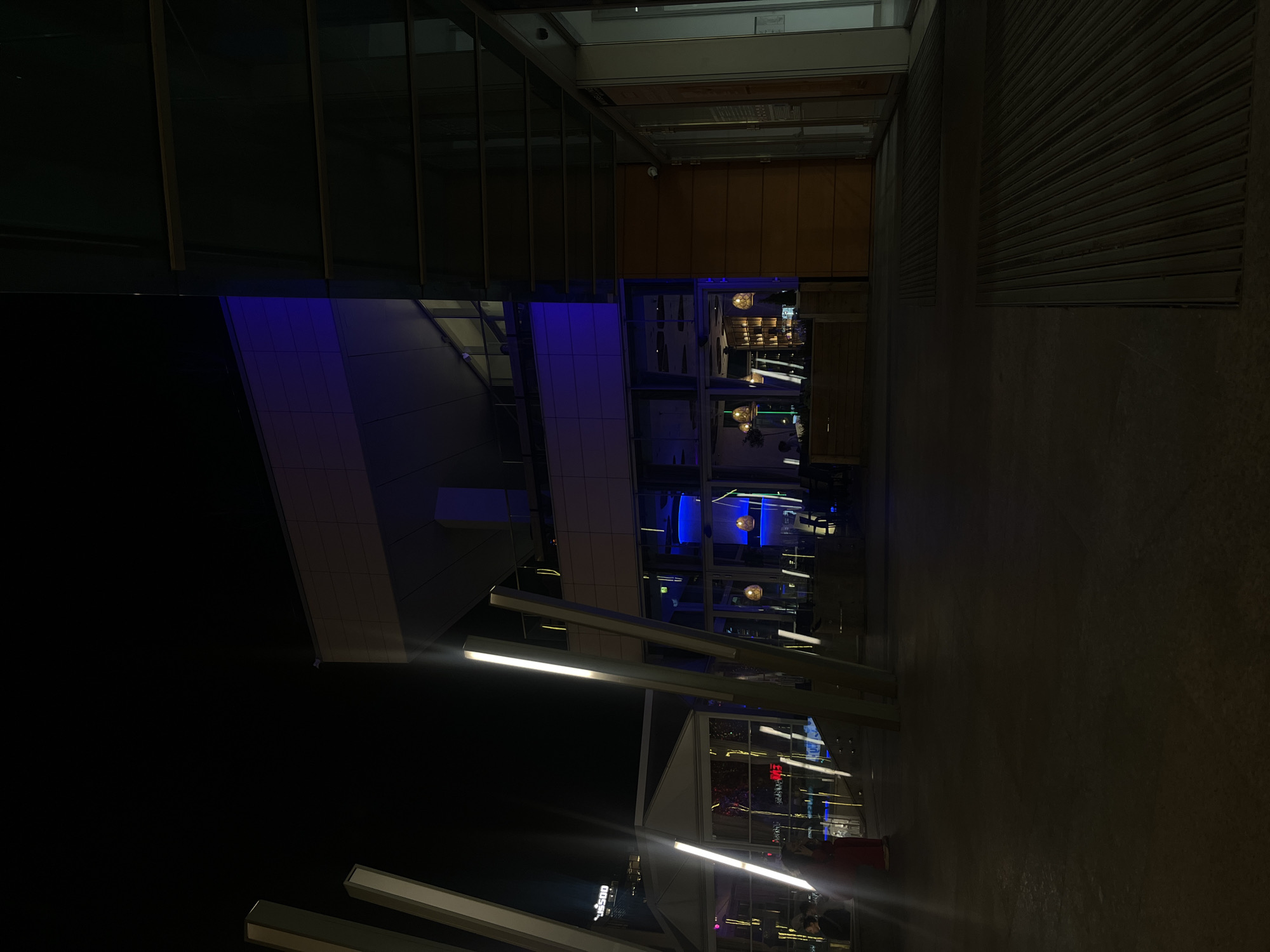 This is a nighttime shot showing a modern building exterior and urban street scene. The building features glass facades, some illuminated with blue lighting. There is a visible restaurant or bar with lights and possible patrons inside. The street is dimly lit, with illuminated light fixtures. I do not have any data to establish the precise location in the city of Sofia, Bulgaria.