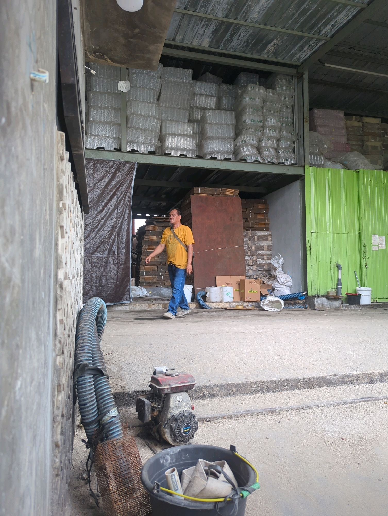 In Batu Belaman, Indonesia, a man in a yellow t-shirt and blue jeans walks through an open-air warehouse. He carries a brown strap over his shoulder. The warehouse has a concrete floor and a metal roof. Stacks of plastic bottles are visible on shelves in the background. To the left, a gray concrete wall with a textured pattern is present. A large, blue corrugated hose snakes along the ground near the wall. In the foreground, a small red and black engine sits on the concrete, next to a black bucket filled with white fabric and yellow strapping. The scene suggests an industrial or storage setting.