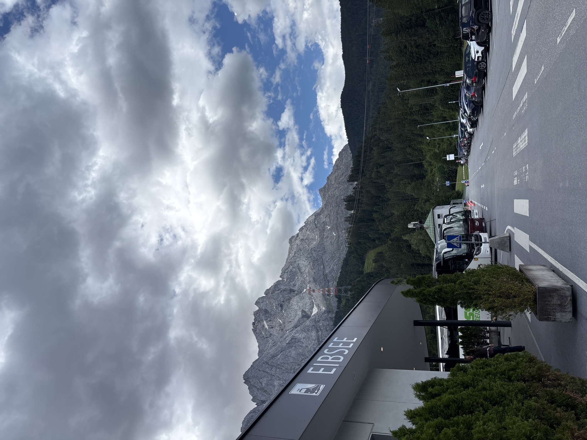 The image shows the Eibsee cable car station in Grainau, Germany, on a cloudy day. A modern building with the name "EIBSEE" prominently displayed in white lettering is on the left. In front of the building, several tour buses are parked, with passengers visible near one of the buses. A parking lot filled with cars is to the right of the buses. In the background, a majestic mountain range dominates the landscape, with a cable car pylon visible against its rocky slopes. Dense green forests cover the lower parts of the mountains. The sky is filled with dramatic grey clouds, interspersed with patches of blue sky. The ground in the foreground is paved asphalt, with white pedestrian crossings.