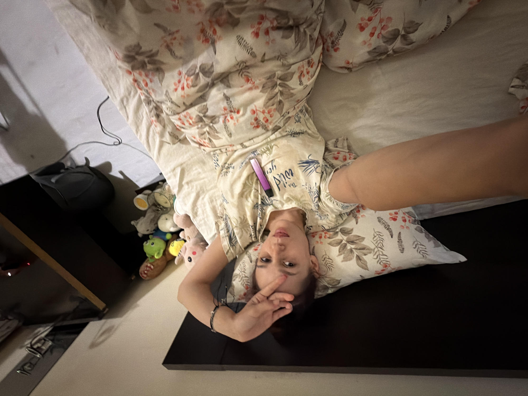 A young woman lies on a bed, holding her hand to her forehead. She is wearing a light-colored t-shirt with a floral and text pattern, and is covered by a blanket with a similar design. A purple object, possibly a vape pen, rests on her chest. To her right are several stuffed animals and a black backpack on the floor. The scene appears to be an indoor setting, likely a bedroom. The presence of a dark wooden headboard suggests the bed is against a wall. The overall lighting is dim, creating shadows.