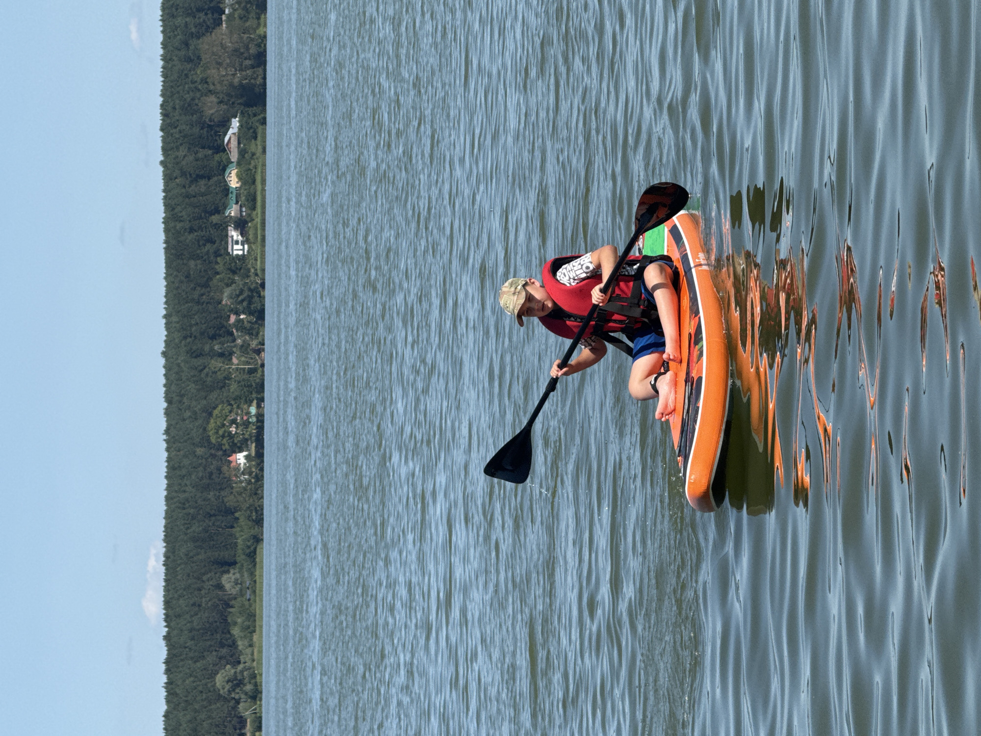A young person is paddleboarding on a calm lake on a sunny day. The person is wearing a red life vest and a camouflage hat, and they are holding a paddle. The paddleboard is orange with some green and black accents. In the background, there is a dense forest line with some houses visible amongst the trees. The sky is clear blue with a few wispy clouds. The water has gentle ripples, and the reflection of the paddleboard and the sky can be seen on the surface. This scene is situated on Lake Votkinsk, which is located in the city of Votkinsk, Russia.