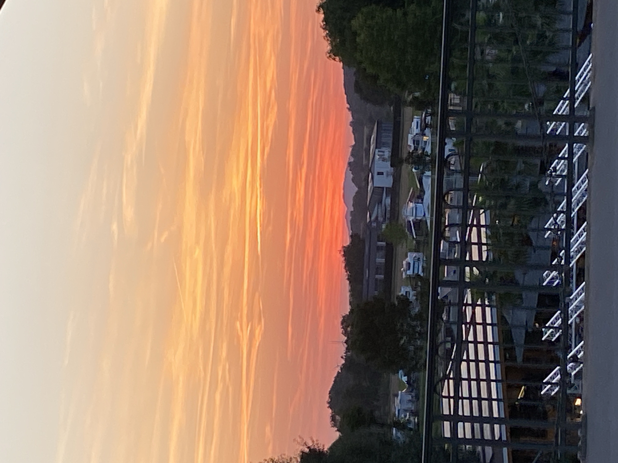 The image shows a sunset over a rural landscape, possibly near Rinteln, Germany. The sky is filled with vibrant orange and pink hues, with thin, wispy clouds stretching across the horizon. In the middle ground, there are several buildings, including what appears to be a campground with several mobile homes and RVs. Trees and rolling hills form the backdrop. In the foreground, a dark metal railing is visible, suggesting the photo was taken from a higher vantage point, such as a bridge or an elevated walkway. The overall atmosphere is peaceful and serene, with the warm colors of the sunset casting a tranquil glow over the scene. There are no people or animals visible in the image.