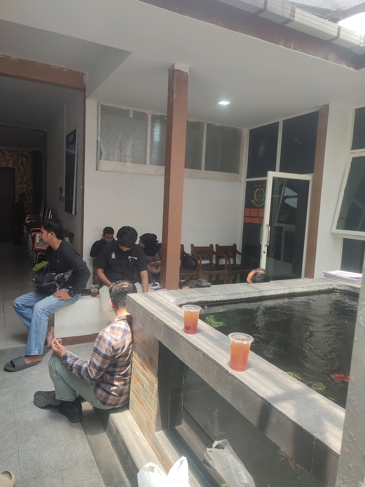 The image depicts an outdoor courtyard in Nganjuk, Indonesia, adjacent to a building with white walls and large windows. Several men are present in the scene. In the foreground, two plastic cups filled with a brown liquid, likely iced tea, sit on the edge of a pond. The pond contains dark water with visible fish swimming within it.
To the left, a man wearing a black long-sleeved shirt, blue jeans, and black sandals is seated on a low concrete ledge, facing away from the camera. He has a black bag slung across his chest. Next to him, another man in a plaid shirt and light-colored pants is sitting on a step, leaning against a tiled wall. He is looking down.
Further back in the courtyard, several other men are seated on similar ledges. One man in a black uniform shirt with a badge is looking down. Another man in a black shirt and pants is seated behind him. A person in a black headscarf is also visible in this group.
The environment appears to be a gathering area, possibly a waiting area or a place for casual interaction. The architecture suggests a relatively modern building, and the presence of the pond adds a decorative element to the space. The lighting indicates it is daytime, with sunlight casting shadows. The background shows a doorway with a glass door, a logo with green and gold elements, and several wooden chairs.