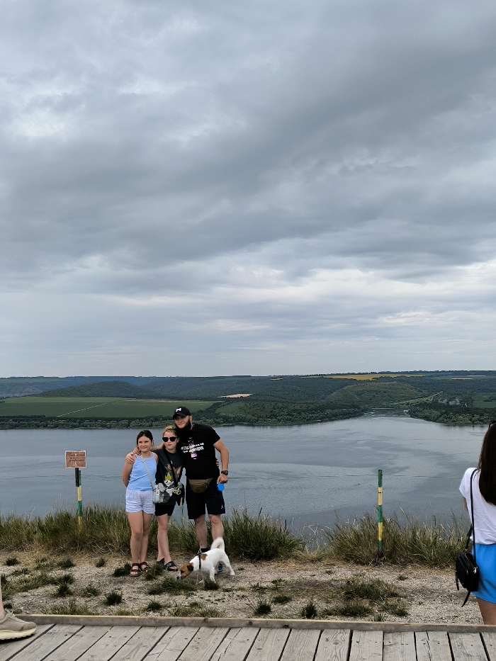 This image depicts an outdoor scenic overlook in the area of Horaivka, Ukraine, under an overcast sky. The foreground features a wooden platform transitioning to a dirt and sparse grass area where three individuals and a small dog are positioned. On the left, a brown information sign is mounted on a yellow and green striped pole. From left to right in the central group: a pre-teen female in a light blue tank top and white shorts stands beside a young adult female wearing sunglasses, a dark t-shirt, and dark shorts, carrying a dark shoulder bag. To their right, an adult male with a beard and a dark baseball cap wears a dark t-shirt, dark shorts, and a light-colored fanny pack; his left arm is around the central female's shoulders, and he holds a blue object in his right hand. A small, white and brown Jack Russell Terrier-type dog stands at their feet, appearing to be on a leash. A fourth individual, primarily visible from the back in a white t-shirt and blue shorts, is partially cropped on the far right. The midground is dominated by a wide, calm body of water. The background reveals undulating green hills and forested land, with interspersed patches of lighter green and yellow agricultural fields. The upper third of the image shows a sky completely covered by dense grey clouds.