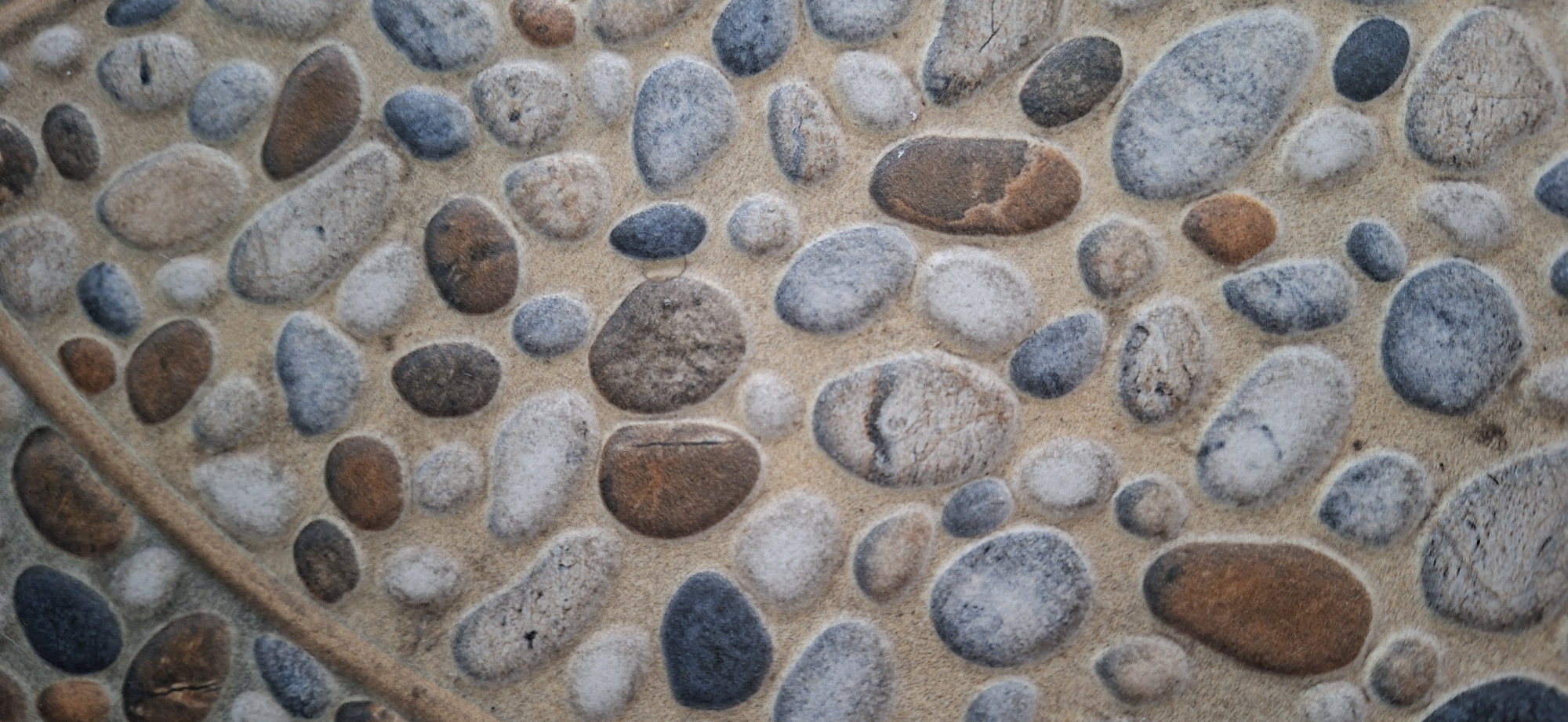 The image displays a close-up, eye-level view of a textured surface resembling a stone pathway or flooring. The surface is composed of numerous smooth, rounded pebbles of varying sizes and colors, including shades of grey, blue, brown, and off-white. These pebbles are embedded in a tan-colored grout or sand-like material, creating a natural, mosaic-like pattern. A curved, lighter-colored line, possibly a vine or a decorative element, runs diagonally across the upper left portion of the image, separating some of the pebbles. The lighting appears to be even, highlighting the textures and subtle variations in the stone colors. There are no people, animals, or prominent landmarks visible in this abstract, patterned composition. This surface could be found in various outdoor or decorative settings within Surabaya, Indonesia, such as a park, garden, or a courtyard.