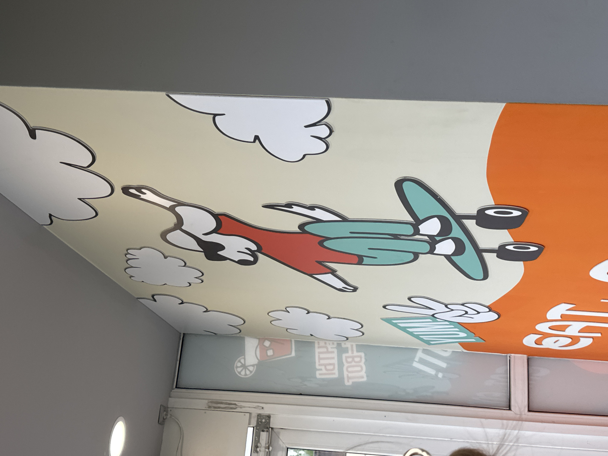 The image shows a colorful mural on a wall, likely in Chaikovsky, Russia, depicting a cartoon dog character skateboarding. The dog is wearing a red shirt, green pants, and sunglasses. It is surrounded by stylized white clouds. In the foreground, the words "YUM!" and "EAT." are visible, also rendered in a cartoonish style with the latter on an orange background. To the left of the mural, a glass door with a reflection of a drink and text is visible, possibly indicating a cafe or restaurant. The lighting suggests an indoor setting with a bright overhead light visible in the upper left corner. The wall is painted a light yellow or cream color, and the mural elements appear to be three-dimensional cutouts.