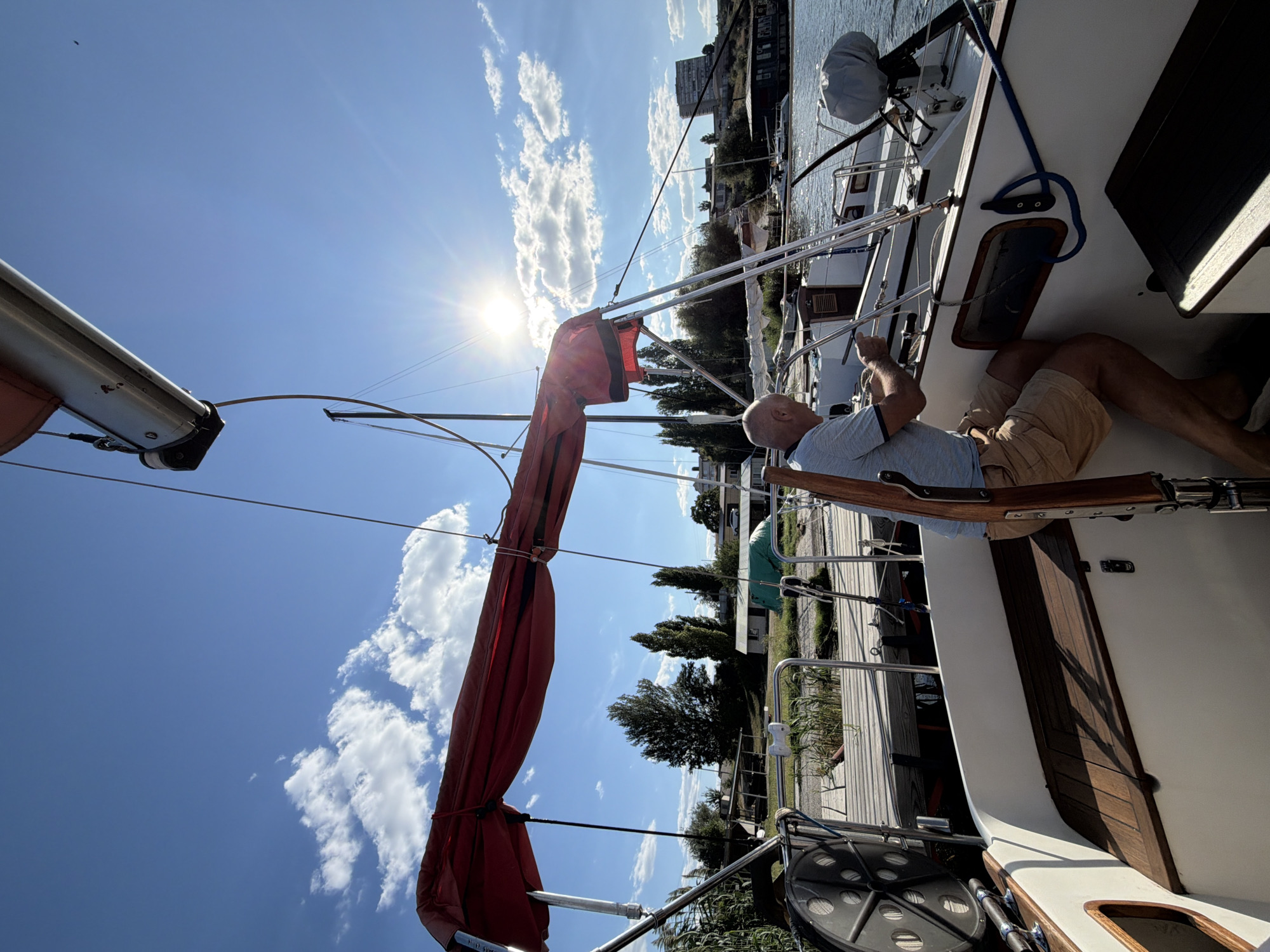 A man is on a sailboat on a sunny day. The man is wearing a blue and white striped polo shirt and tan shorts. He is standing and holding onto a steering wheel. The sailboat has a red awning over the cockpit. In the background, there is a wooden dock, trees, and buildings. The sky is blue with some white clouds. The image appears to have been taken in the city of Kamyshin, Russia, as it is a port city located on the Volga River.