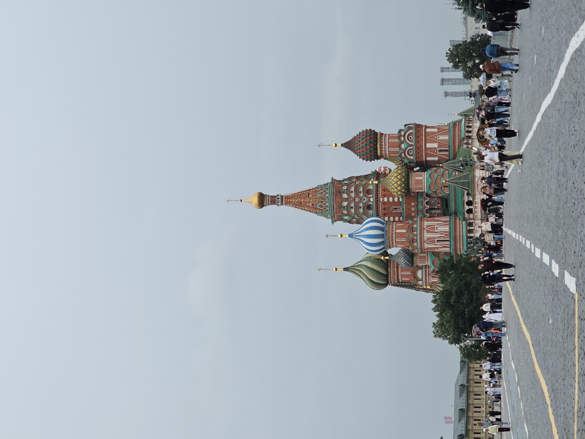 This is a photograph of Saint Basil's Cathedral, located in Red Square, Moscow, Russia. The cathedral is a vibrant and iconic landmark, featuring a collection of vividly colored onion domes of various shapes and patterns. The foreground is a cobblestone plaza with a yellow line and dashed white lines painted on it, indicating pedestrian pathways. Numerous people are walking on the plaza, some carrying umbrellas, suggesting it may be raining or overcast. The sky is a uniform light gray. To the left of the cathedral, a long, low-rise building with a light-colored facade and arched windows is visible. Trees are scattered throughout the scene, and in the background, beyond the cathedral, industrial smokestacks can be seen.