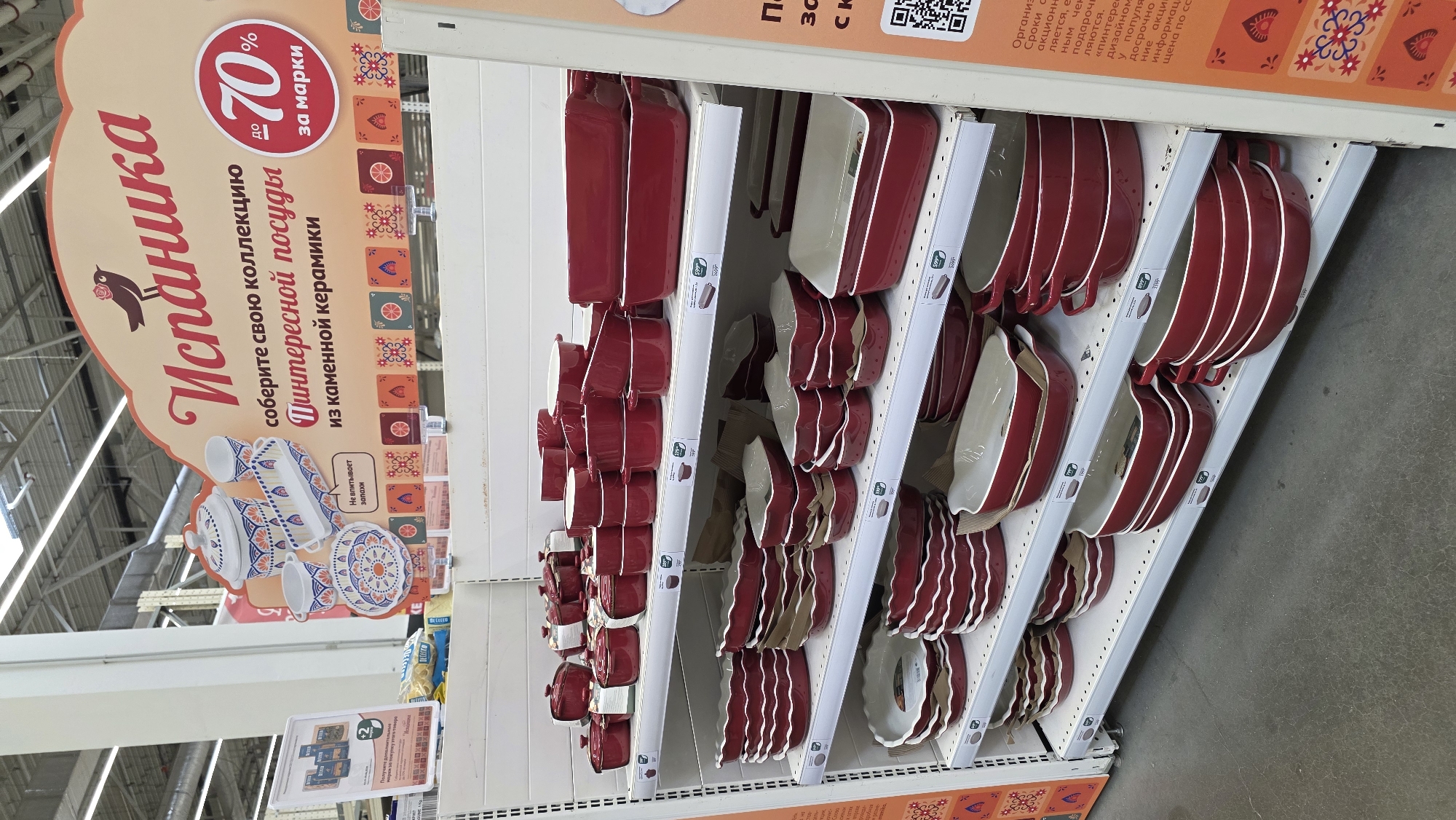 The image captures an aisle within a retail store, likely a supermarket or home goods store, showcasing a collection of ceramic kitchenware. Numerous red and white baking dishes, casseroles, and other oven-safe ceramic items are neatly stacked on multiple white shelves, presented for sale. The dishes feature a distinctive red exterior with a contrasting white interior and rim, appearing in various rectangular, oval, and round shapes, some with integrated handles.
A large, decorative hanging sign above the display features the brand name "Испаника" (Ispanika) alongside a small bird graphic. The sign encourages customers to "collect your collection of interesting dishes made of stoneware ceramics." A prominent circular element advertises a discount "up to 70% for loyalty stamps/points." Smaller text on the sign notes that the products "do not absorb odors."
To the right, a partially visible vertical sign outlines the terms and conditions of the promotion, mentioning details such as "promotional dates" and "information." Smaller price tags or promotional labels are visible on the shelves among the products, some indicating potential prices such as "599" or "799." The scene is brightly lit, indicative of an indoor retail setting, with no people visible.