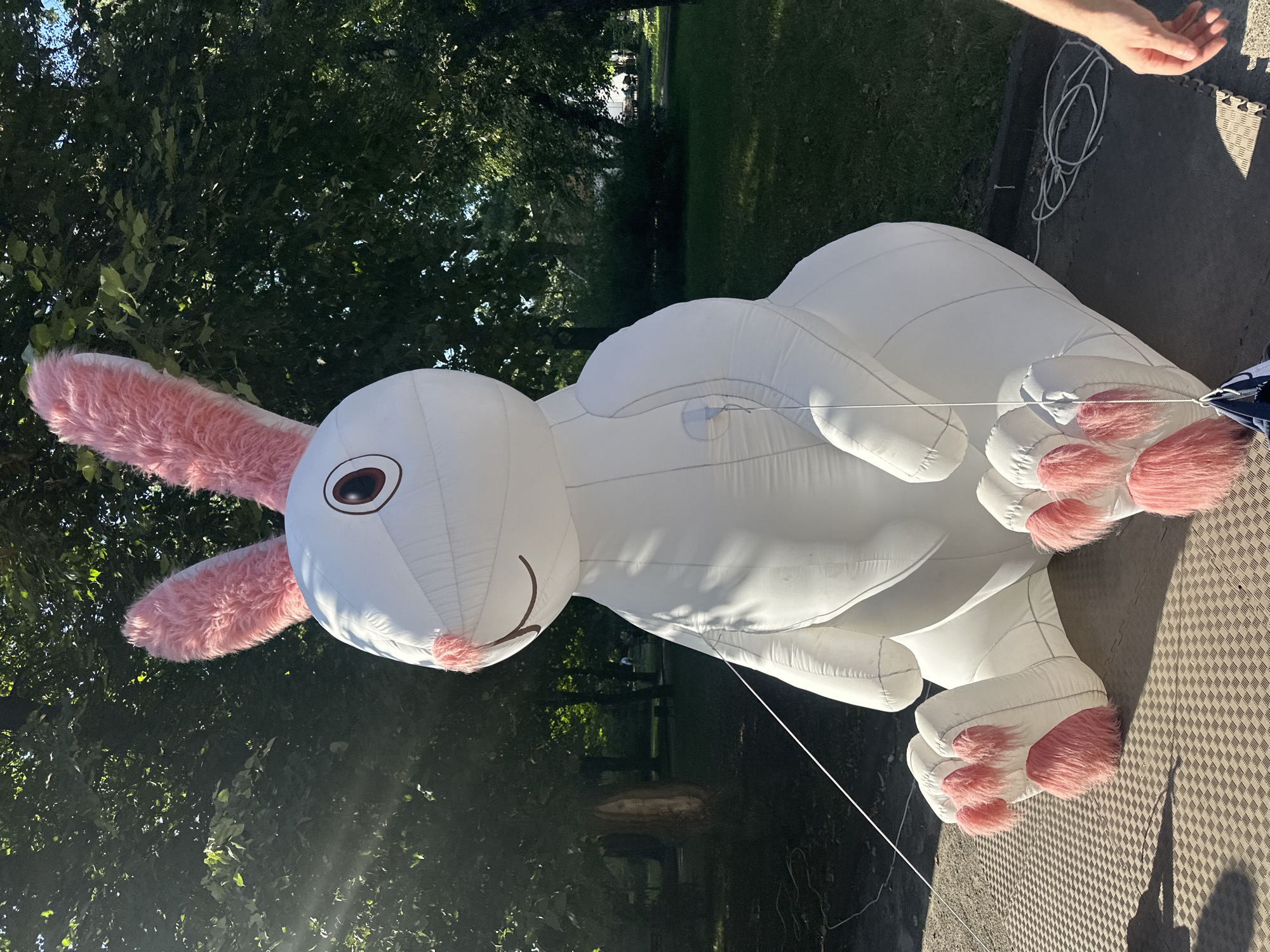 A large, white, inflatable rabbit with pink fuzzy ears and paw pads is sitting on a grey, textured mat outdoors. The rabbit has a simple face with brown eyes and a drawn-on smile and nose. It is surrounded by lush green trees, with sunlight filtering through the leaves. In the background, a park area with grass and distant figures can be seen. A person's hand is visible in the lower right corner of the image. The scene appears to be set up in a public park, possibly for an event. The inscription "Ternopil, Ukraine" is provided as context for the location.