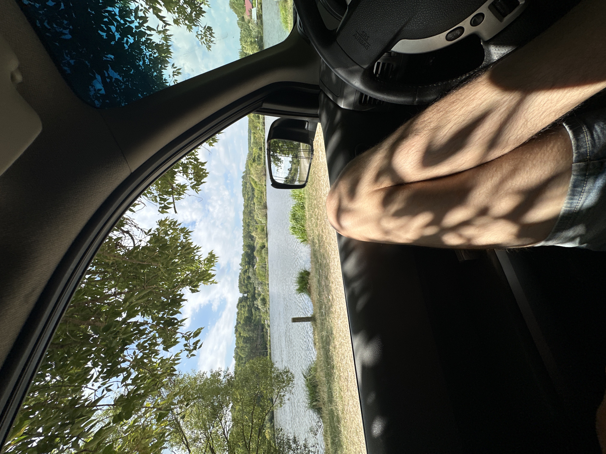 The image shows a view from inside a car, looking out the passenger side window towards a lake. The foreground is dominated by the interior of the car, including the door panel and a portion of a steering wheel. A person's legs, clad in denim shorts, are visible crossing in front of the window. Sunlight casts shadows of tree branches across the legs and the car's interior. Outside the car, a body of water, likely a lake or river, stretches across the midground, with a vegetated shoreline and rolling hills covered in trees in the background. The sky is blue with scattered white clouds. The scene appears to be a natural landscape. There is no explicit information in the image to indicate the specific location in Makiv, Ukraine.