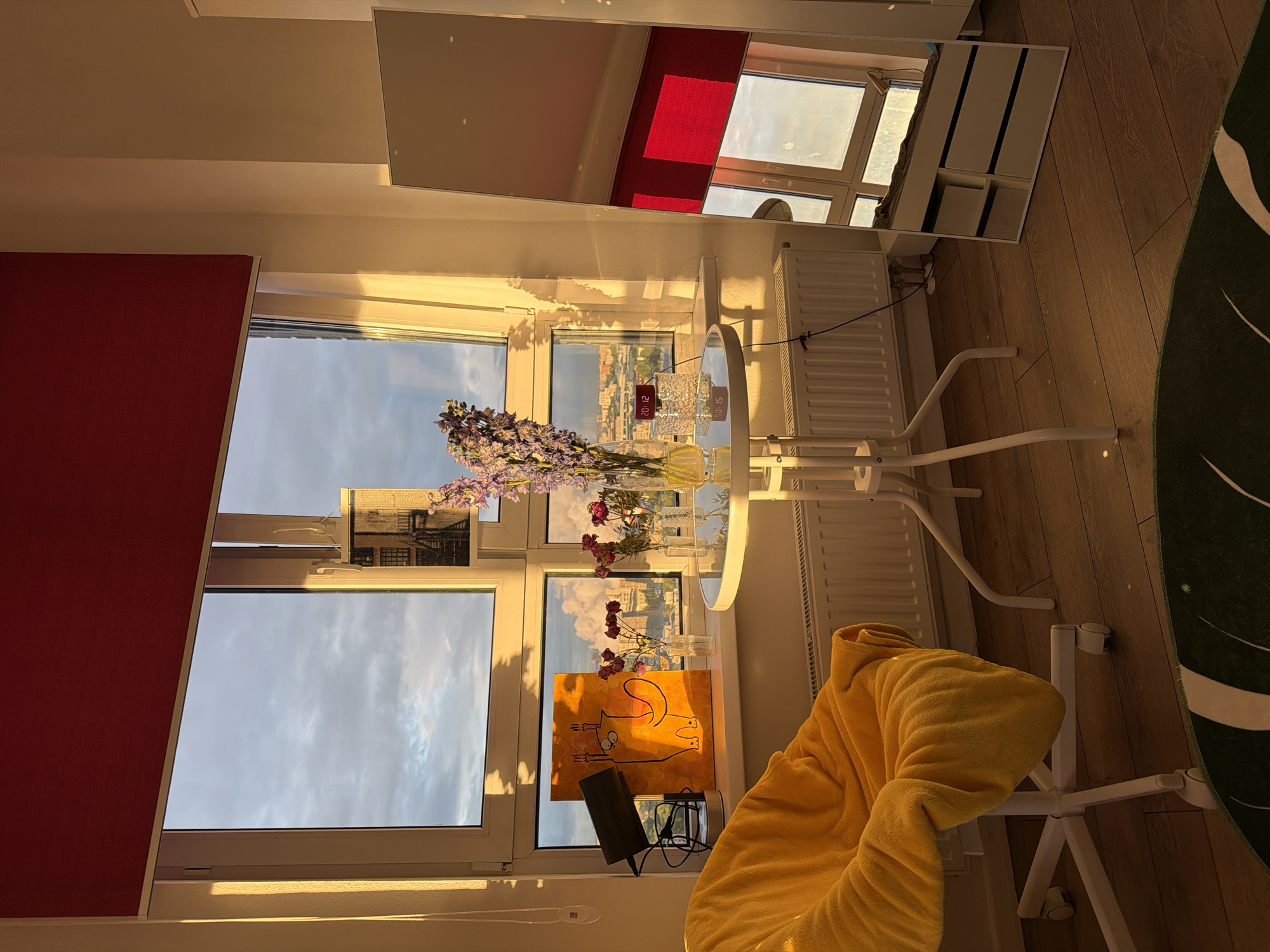 The image depicts an interior scene with warm lighting. A window with a red blind is visible, showcasing a cityscape beyond. On the windowsill, there are flowers in vases, a painting, and various objects. A round table is positioned in front of the window, and a chair with a yellow blanket is nearby. A mirror reflecting the room and window is also present. The room has wooden flooring and a white radiator.