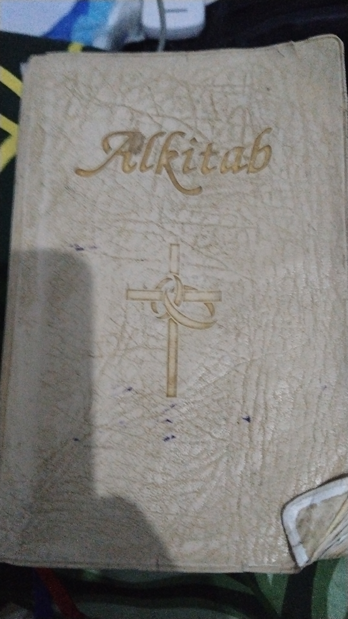 A light-colored, embossed book with the word "Alkitab" in gold script is centered on a dark, textured surface. Below the text is an embossed image of a cross intertwined with a ribbon-like design. The book appears to be slightly worn, with some faint scuff marks and smudges visible on its cover. The background is out of focus, but appears to be a mix of dark and colorful patterns. The image was taken in Banjarsari, Indonesia.