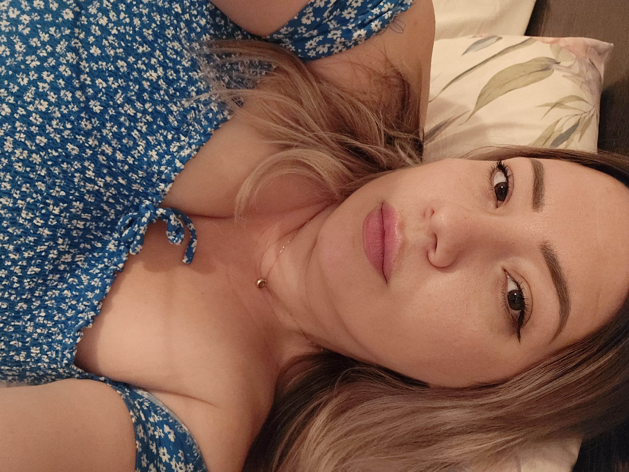 A selfie of a woman lying on a bed with a floral pillow, taken from a slightly elevated angle. She has light brown hair with blonde highlights and is wearing a blue off-the-shoulder top with a white floral pattern. Her makeup includes eyeliner and pink lipstick. A delicate gold necklace with a small circular pendant rests on her décolletage. The scene is indoors, suggesting a bedroom setting. No information about the location in Karlstadt, Germany, can be determined from the image itself.