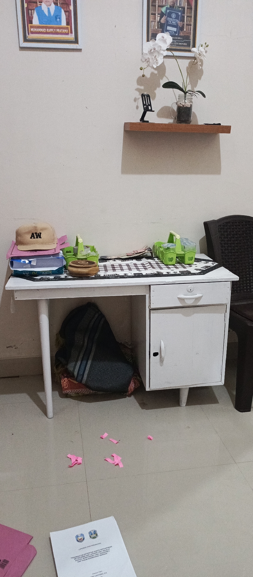The image depicts an indoor scene, possibly a room in Jatisari, Indonesia, characterized by a modest desk and its surroundings. The desk itself is a white, three-legged structure with a drawer and a cabinet on the right side. The top surface of the desk is covered with a checkered cloth, upon which several items are placed. These include a beige baseball cap with the letters "AW" on it, a stack of books, two green plastic baskets containing what appear to be small cups or containers, and a wooden circular object. To the left of the desk, under the surface, a folded patterned cloth is visible.
On the wall behind the desk, two framed pictures are mounted. The left frame contains a portrait of a person in formal attire, with text below that reads "MUHAMMAD RAFFLY PRATAMA". The right frame also contains a portrait of a person, this time in academic regalia, holding a certificate. A wooden shelf is mounted on the wall above and between the pictures, holding a small potted plant with white artificial flowers and a black phone stand.
The floor is tiled in a neutral color. Several small, crumpled pink pieces of paper are scattered on the floor in front of the desk, and a white document with Indonesian text is also visible on the floor. To the right of the desk, a portion of a dark-colored chair is visible. The lighting in the room appears to be artificial and somewhat dim, casting shadows from the objects on the shelf.