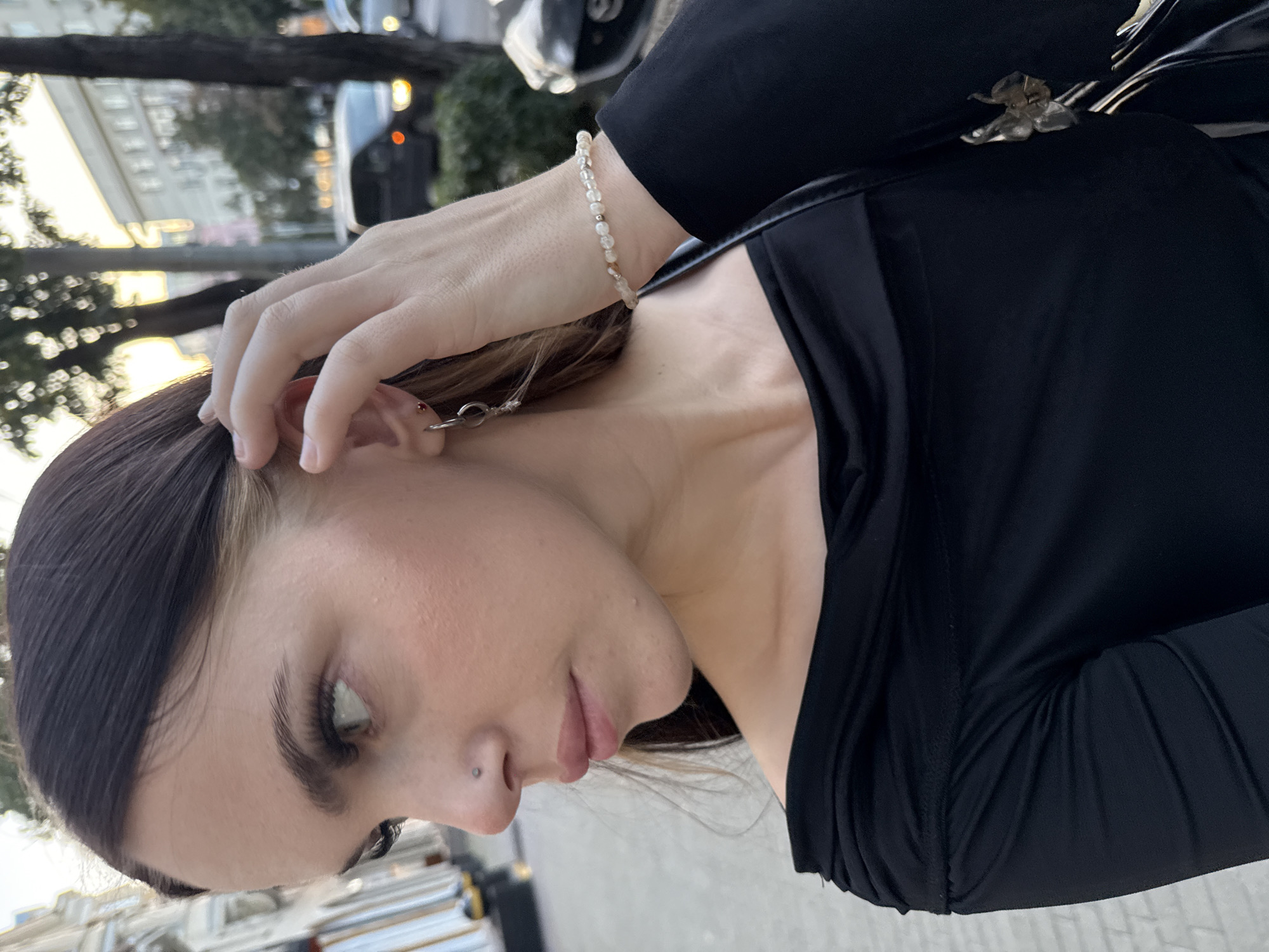 In an outdoor urban environment, a young woman with dark hair, a nose piercing, and eye makeup is captured in a close-up shot. She is wearing a black off-the-shoulder top and a beaded bracelet. Her right hand is raised, touching her hair near her ear, where she is wearing a small hoop earring. The background is blurred, showing trees and buildings, suggesting a street or park setting. The lighting indicates it is likely late afternoon or early evening, with the sun casting a warm glow. This scene is taking place in the city of Chelyabinsk, Russia.
