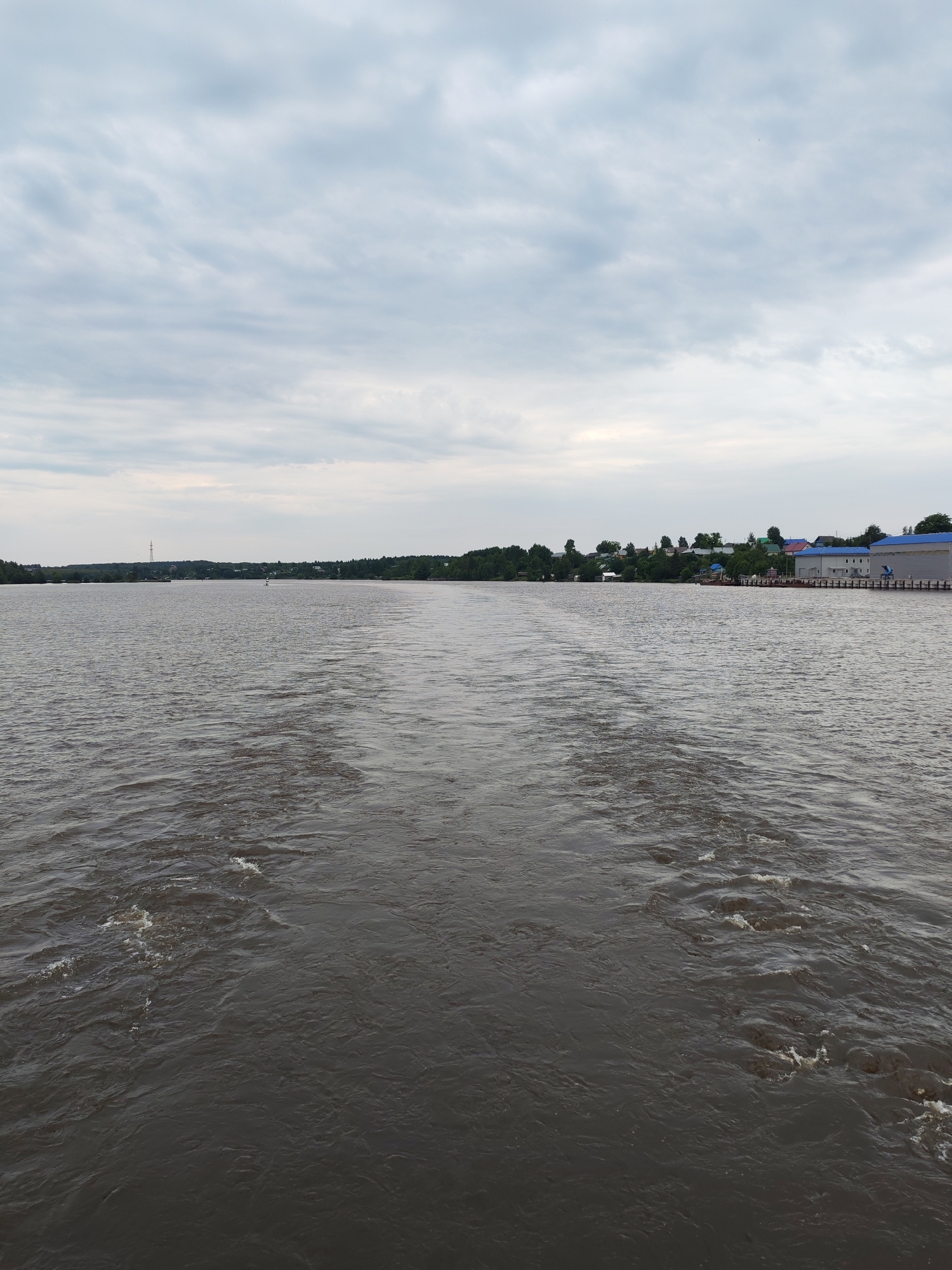 A wide, dark river flows under a cloudy sky. The camera is positioned on the water, looking towards a distant shoreline lined with trees and buildings. In the foreground, a wake fans out behind the viewer, indicating movement through the water. The shoreline on the right side of the image features a cluster of buildings, some with blue roofs, and a pier. On the left side of the distant shore, a tall pylon is visible. The overall impression is a view from a moving vessel on a river in a relatively undeveloped area. This scene is likely located in the city of Ivanov Bor, Russia, given the context.