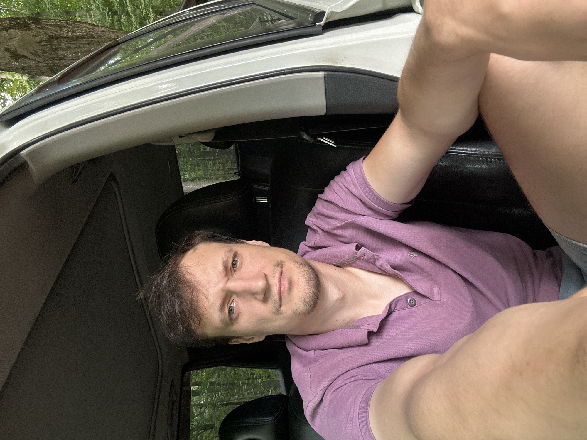 A man with short, dark hair and light skin is shown from the chest up, seated in the passenger seat of a car. He is wearing a short-sleeved, purple polo shirt. The car's interior is visible, including the black leather seats, the headliner, and the white exterior of the car. Through the open car door and windows, a wooded area with green trees is visible, suggesting an outdoor, possibly rural, location. The man's arms are extended forward in the frame. The location is Shatava, Ukraine.