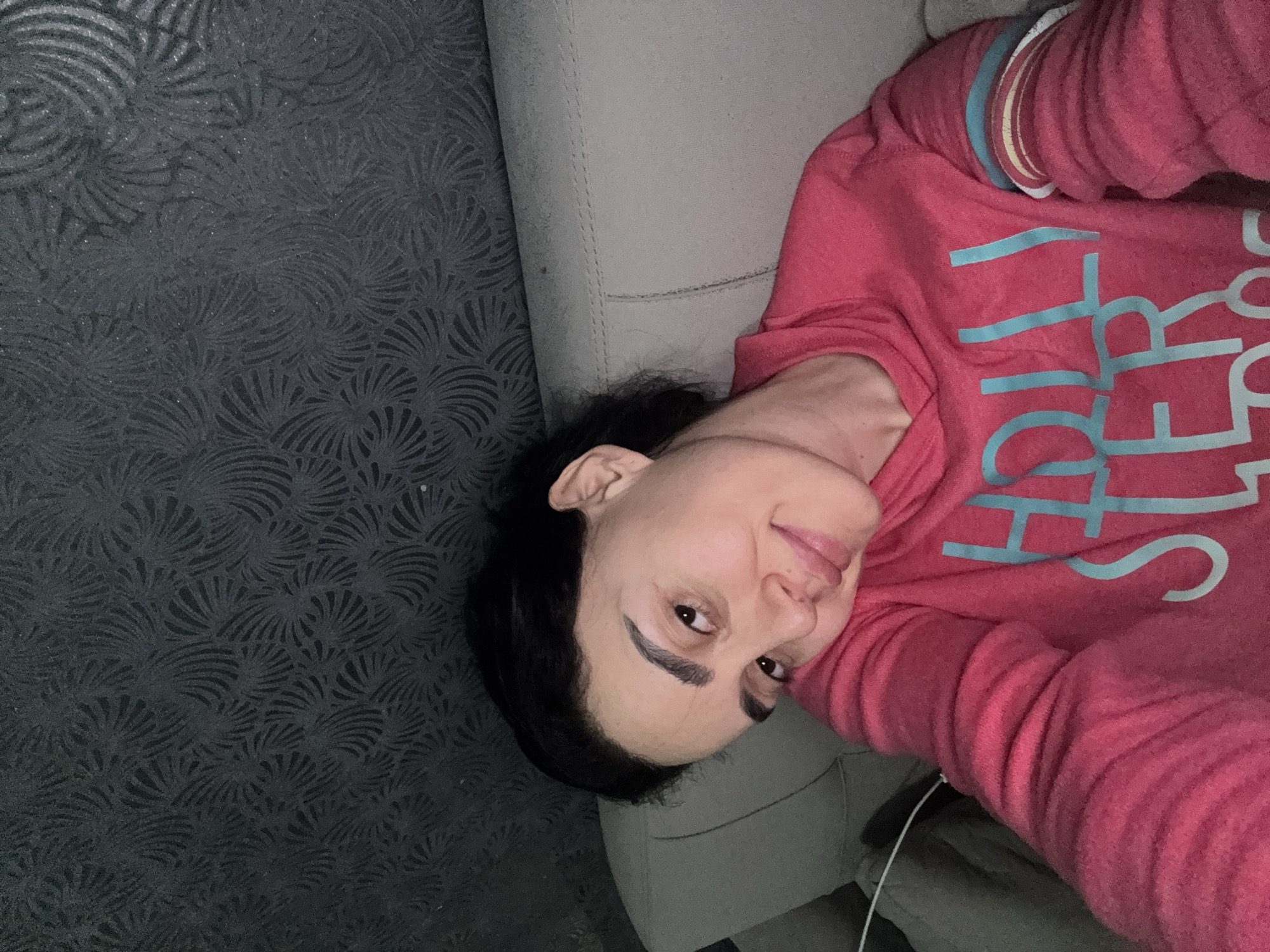 A woman lies on a light gray couch in front of a patterned dark gray wall. She is wearing a pink long-sleeve shirt with light blue lettering. Her dark hair is loose, and she is looking at the camera with a slight smile. The location is not discernible from the image.