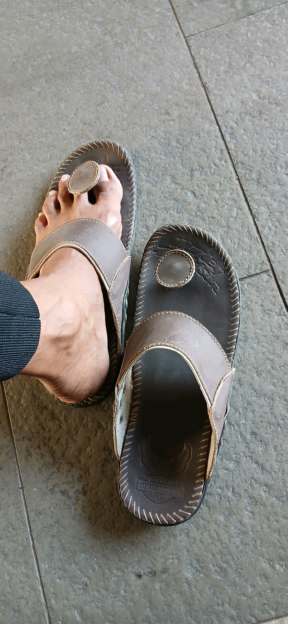 Two brown leather thong sandals are on a tiled floor. One sandal is worn by a person, showing their bare foot and toes. The other sandal is empty. The worn sandal has a dark brown sole and a lighter brown strap that goes over the top of the foot and between the big toe and second toe. The empty sandal also has a dark brown sole with stitching around the edge and a similar lighter brown strap. The sole of the empty sandal has an embossed logo that appears to be a Harley-Davidson emblem. The floor is made of large, rectangular gray tiles with visible grout lines. The overall lighting suggests an indoor or covered outdoor setting.